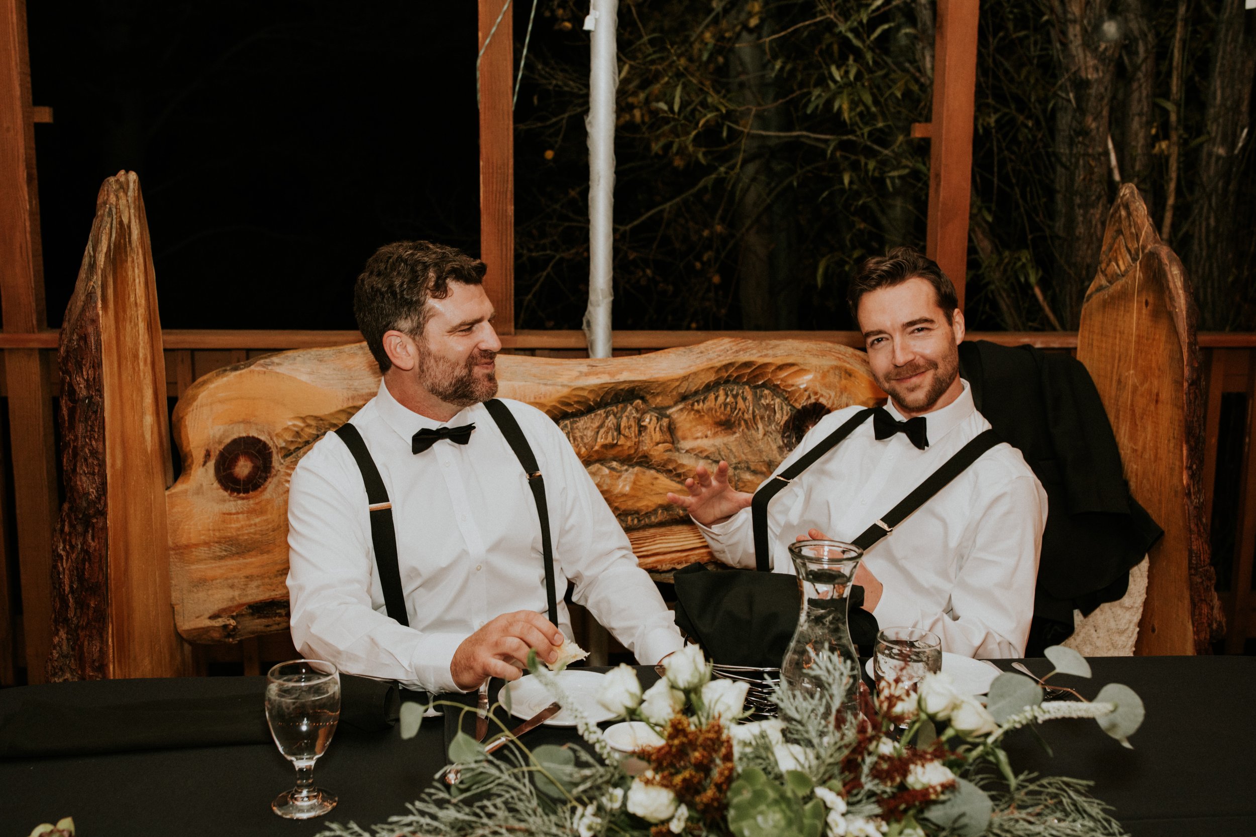Two handsome, gay male grooms in tuxedos with suspenders and bow ties sitting at a formal dinner table at their wedding by Dorey Kronick