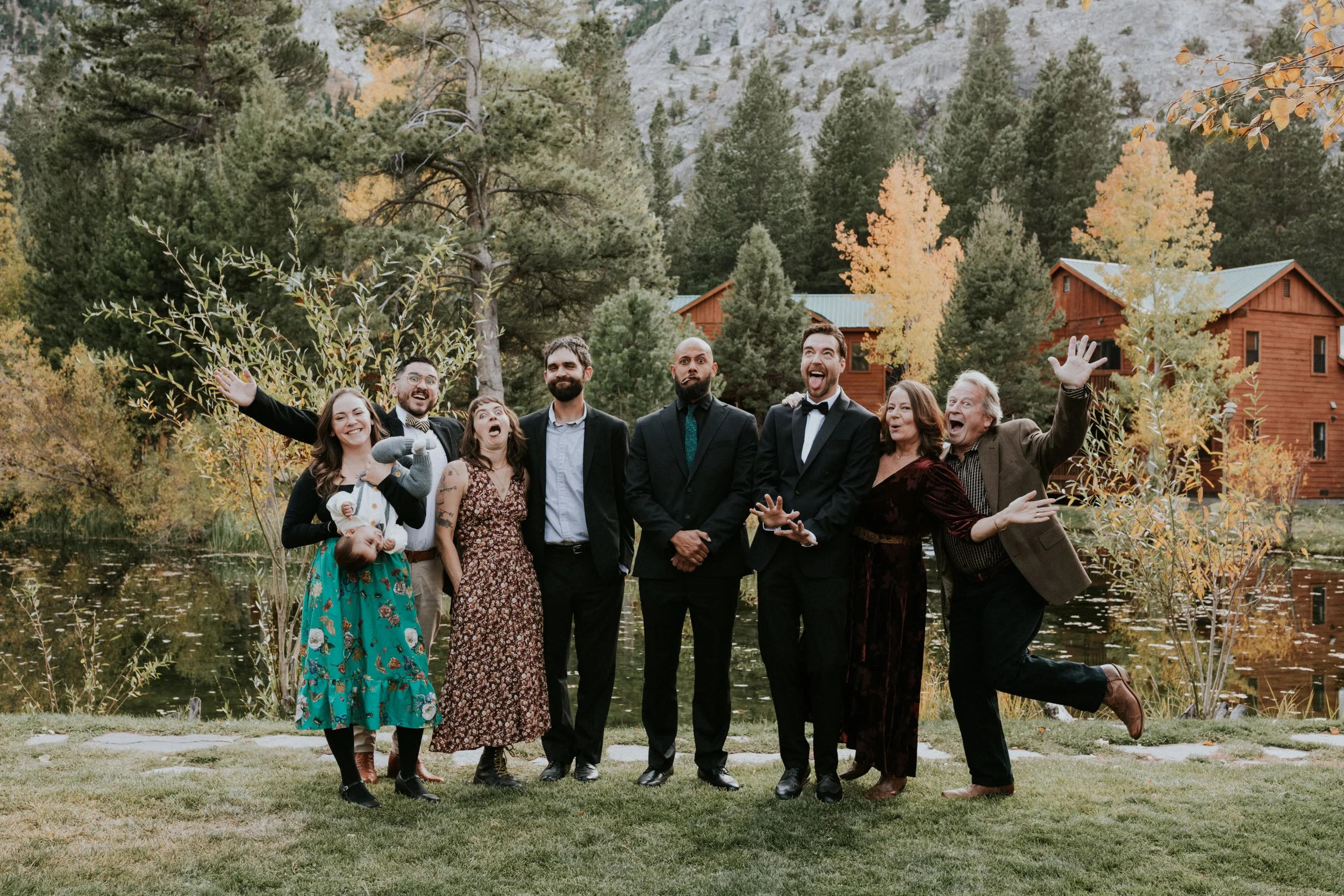Group of cute people standing outdoors near a pond with trees and cabins in the background, celebrating and smiling at Taylor and Dan's wedding in CA by Dorey Kronick