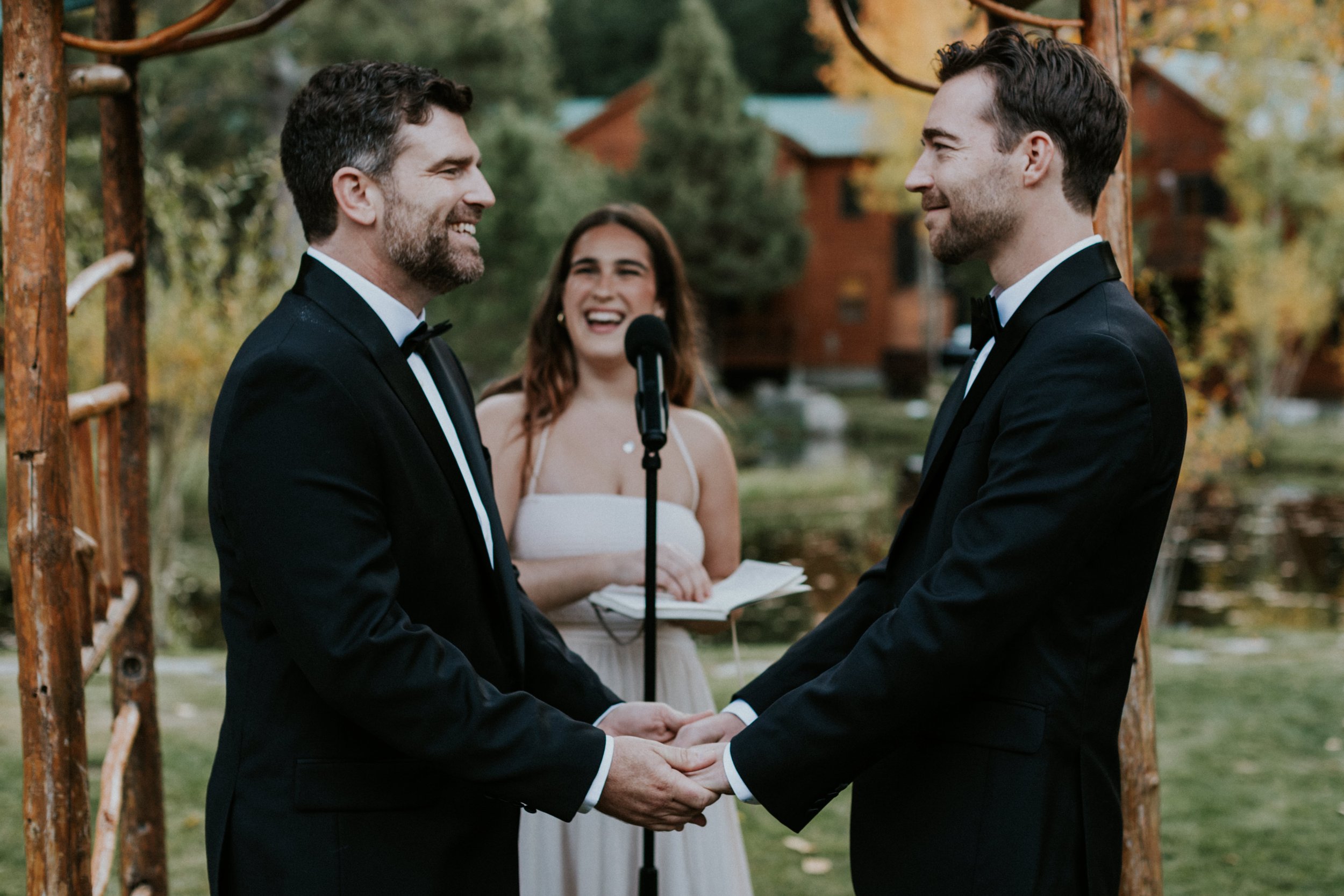 Two gay men in tuxedos holding hands, gazing at each other during their wedding ceremony outdoors, with a woman officiant smiling, under a wooden arch surrounded by trees and greenery at Double Eagle Resort in Mammoth, CA by Dorey Kronick