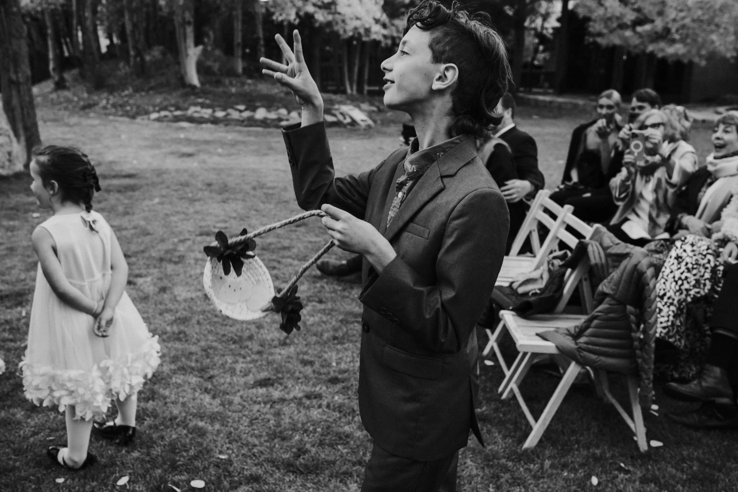 A young man in a suit holding a wand and a paper fan, at an outdoor event, with children and adults seated on chairs behind him, trees in the background, in black and white by Dorey Kronick