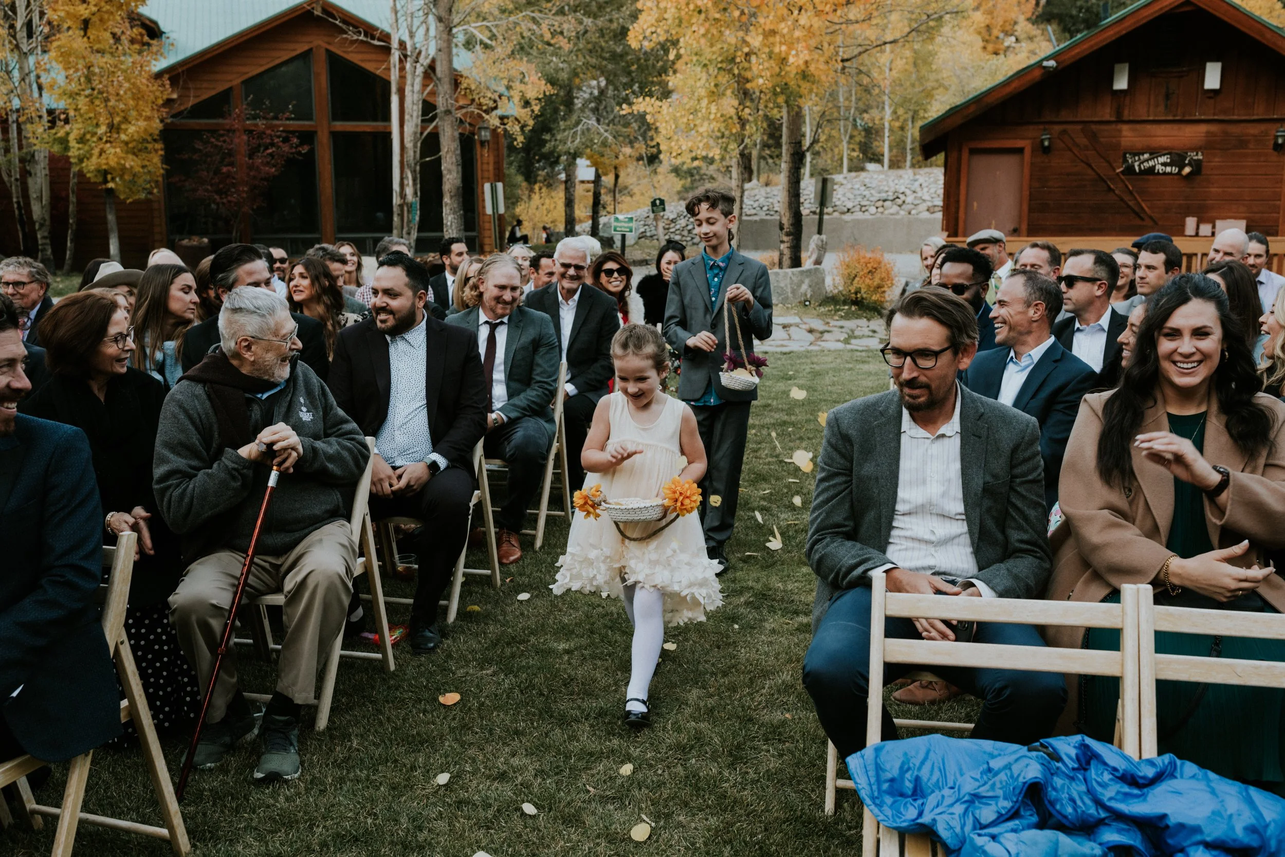 Children walking down the aisle at an outdoor wedding ceremony, surrounded by seated adult guests smiling and laughing, with trees and wooden buildings in the background in June Lake, CA by Dorey Kronick