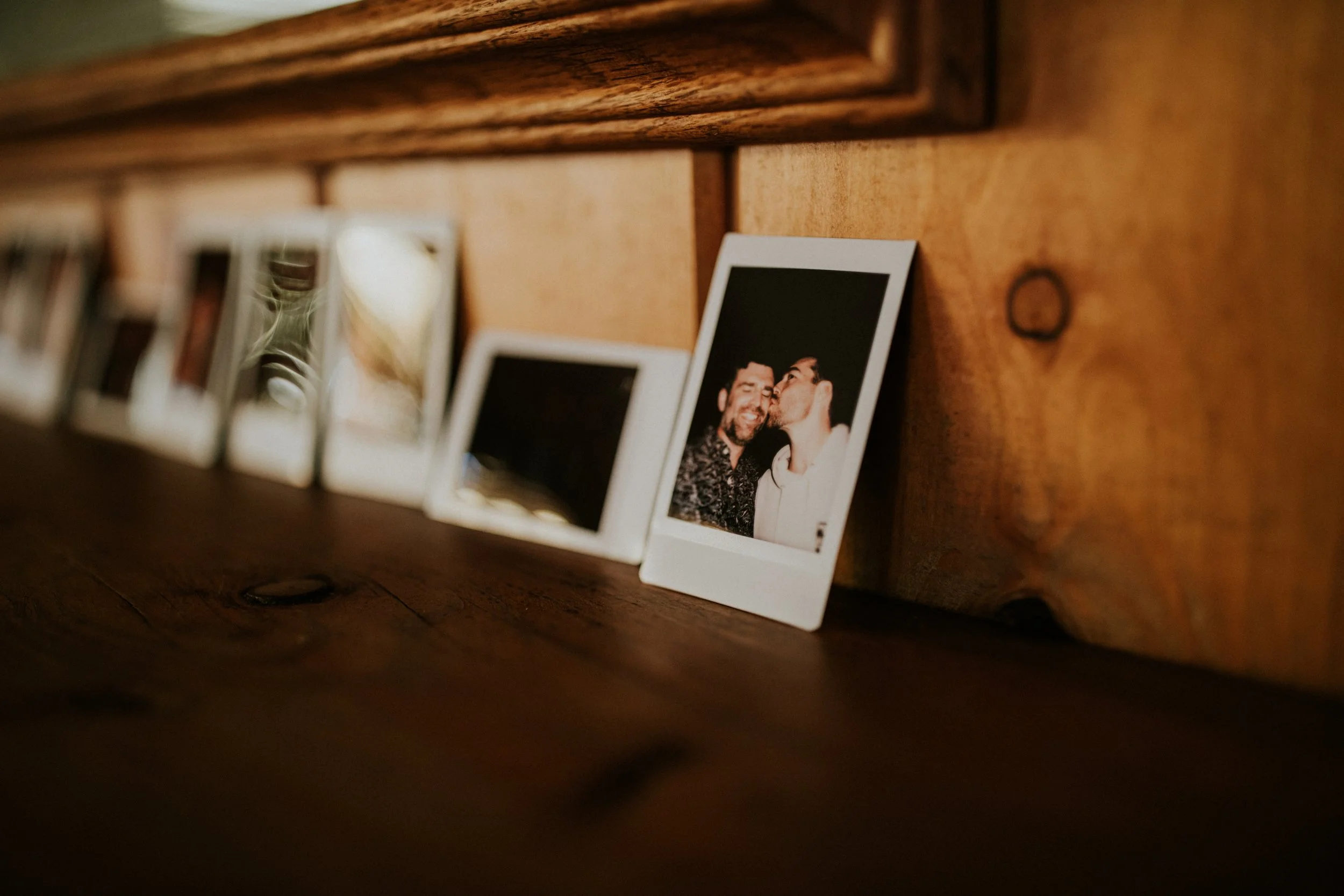 Series of Polaroid photos lined up on a wooden surface leaning against a wooden wall, with the focus on the last photo showing two men, one kissing the other's cheek, both smiling, in June Lake, CA by Dorey Kronick