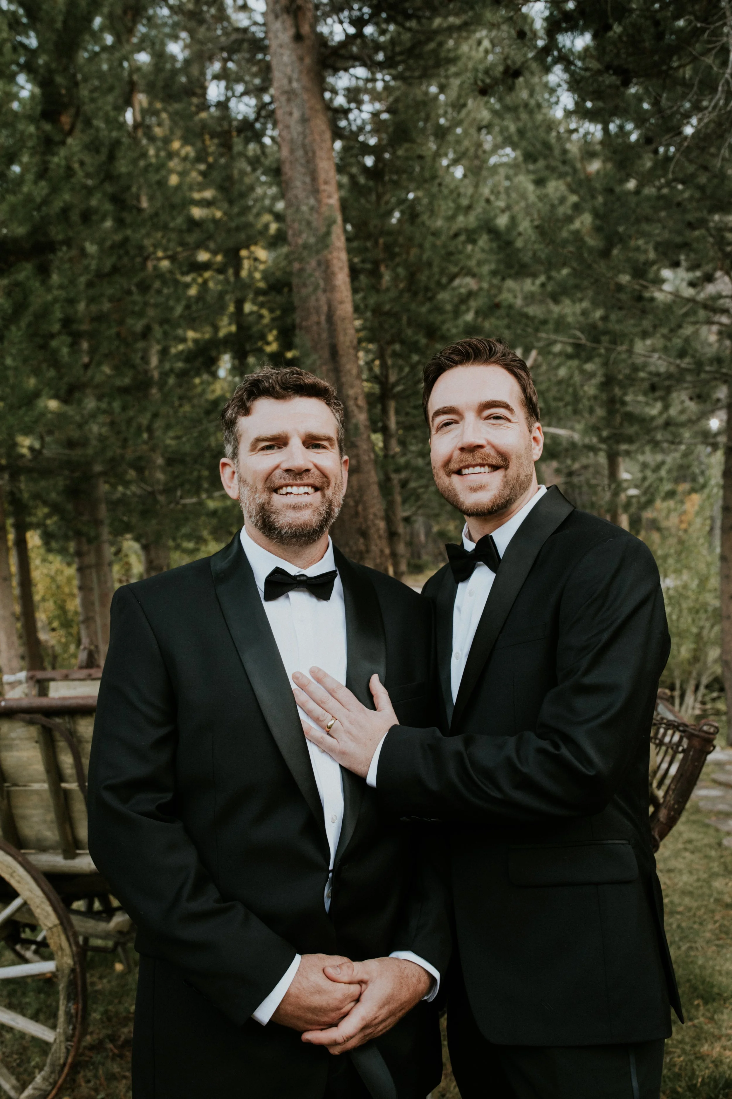 Two handsome gays in tuxedos smiling outdoors, one with hand on the other's chest, surrounded by trees in Mammoth, CA by Dorey Kronick