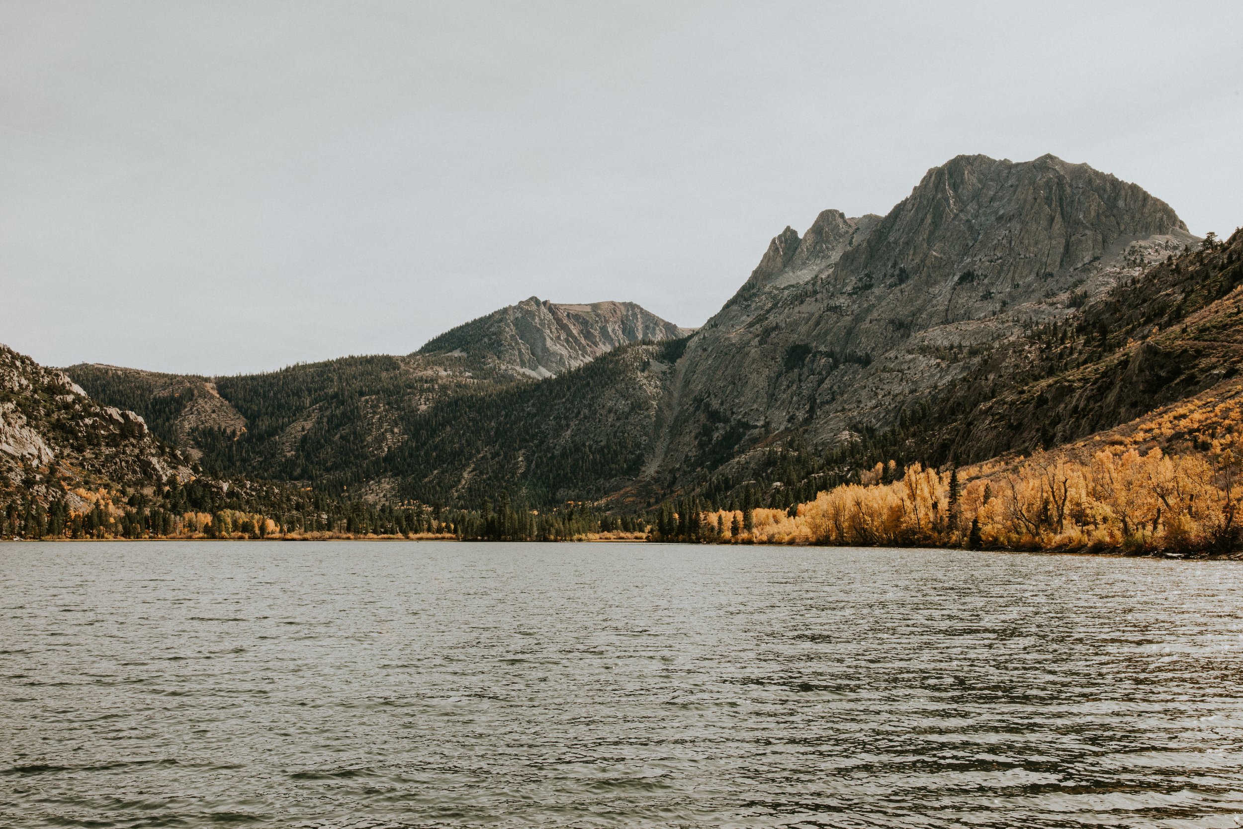 Scenic view of a lake with rippling water, surrounded by forested hills and mountains with rocky slopes, under a cloudy sky in June Lake, CA by Dorey Kronick