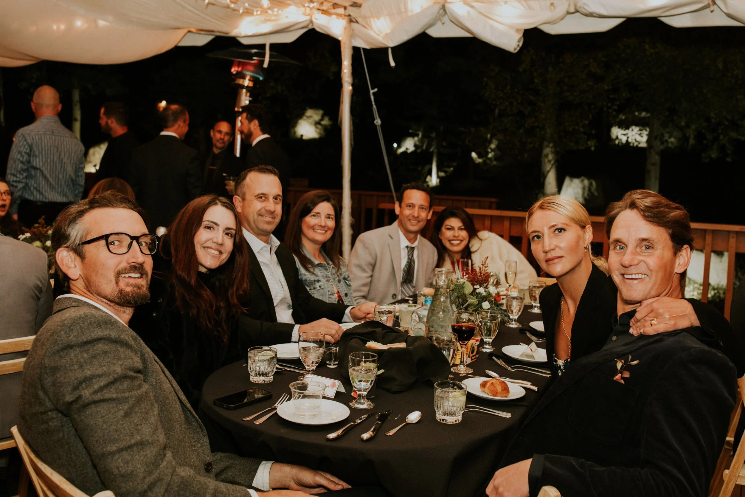Group of happy people sitting at a dinner table, smiling, at an outdoor wedding during the evening by Dorey Kronick