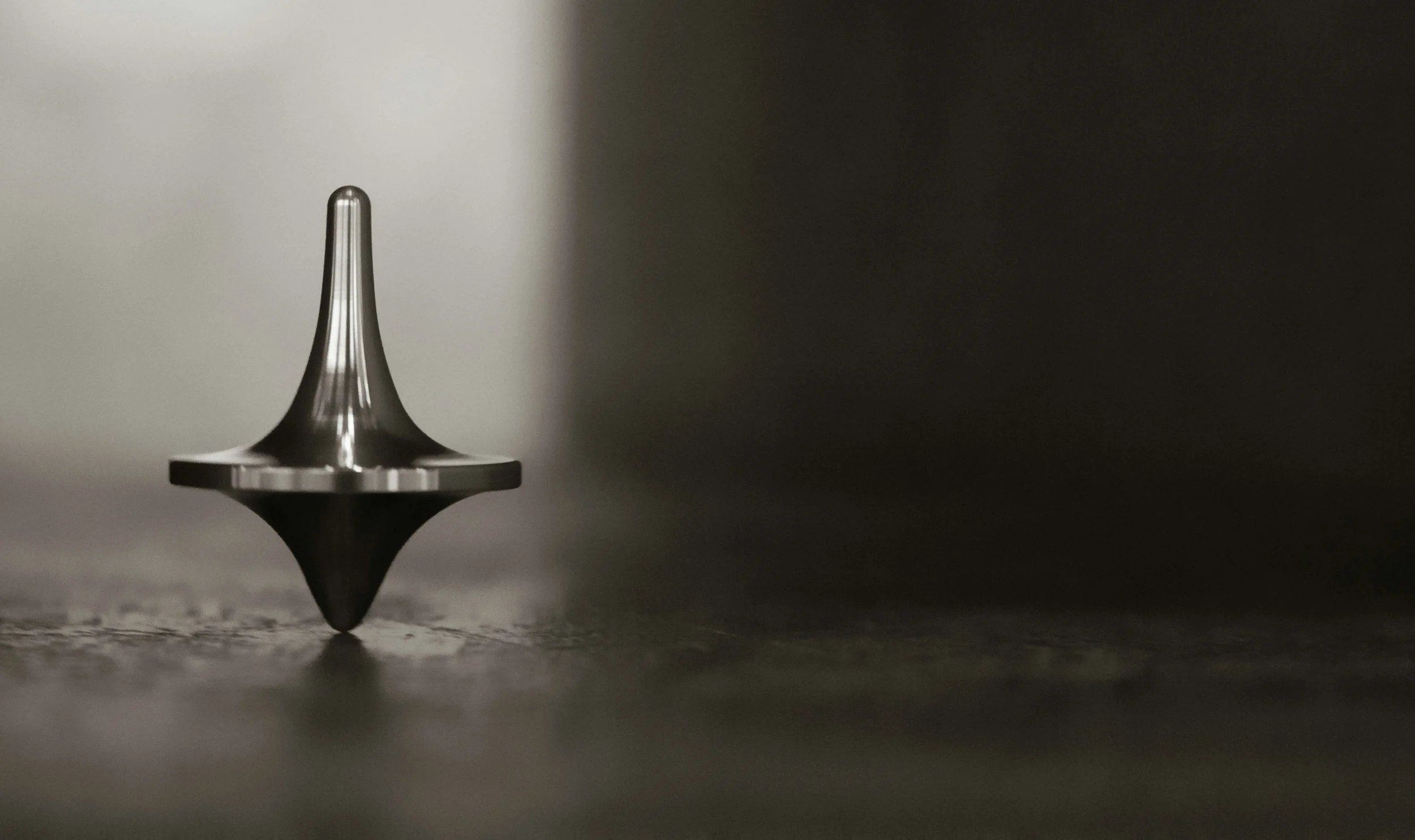 A stainless steel spinning top, spinning upright on a rough surface in the foreground, on a moody, dark and blurred background.