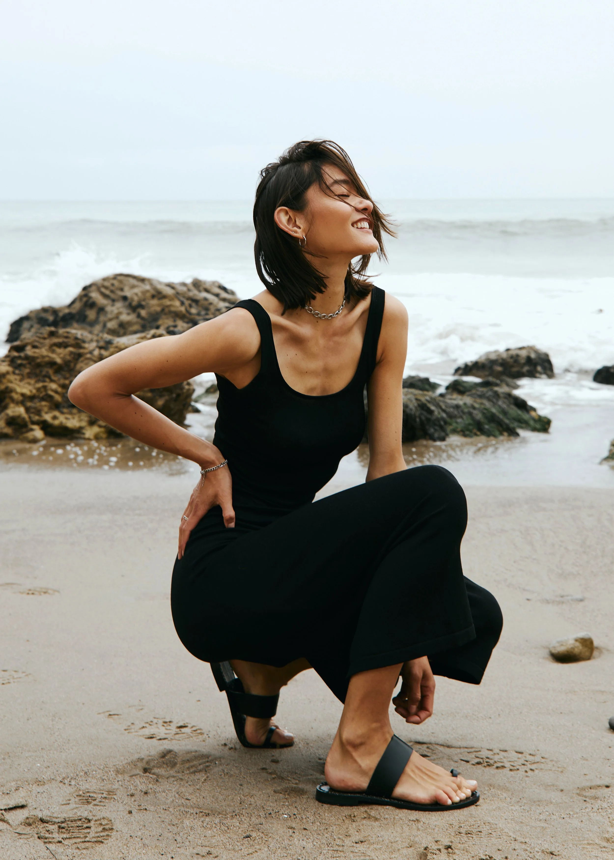 Woman in black tank top and black skirt sitting on sandy beach with ocean waves and rocks behind her, smiling with eyes closed, enjoying the breeze.