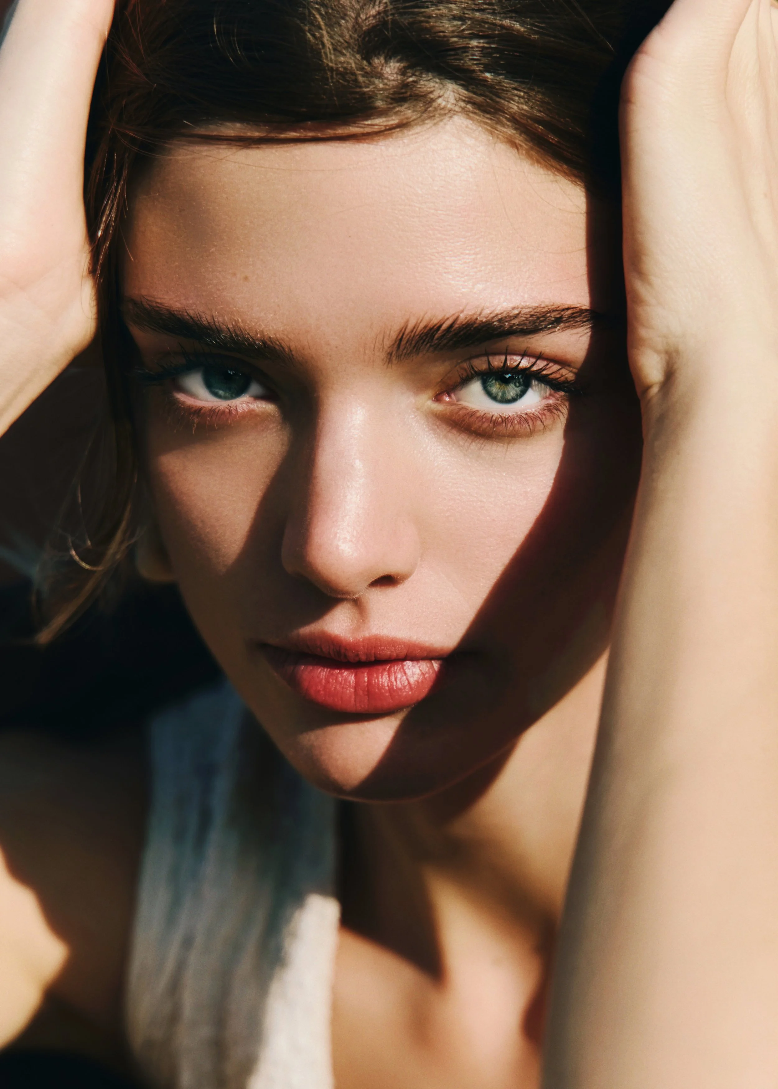 Close-up of a woman's face with blue eyes and fair skin, holding her hair back with her hands, sunlight illuminating her face.