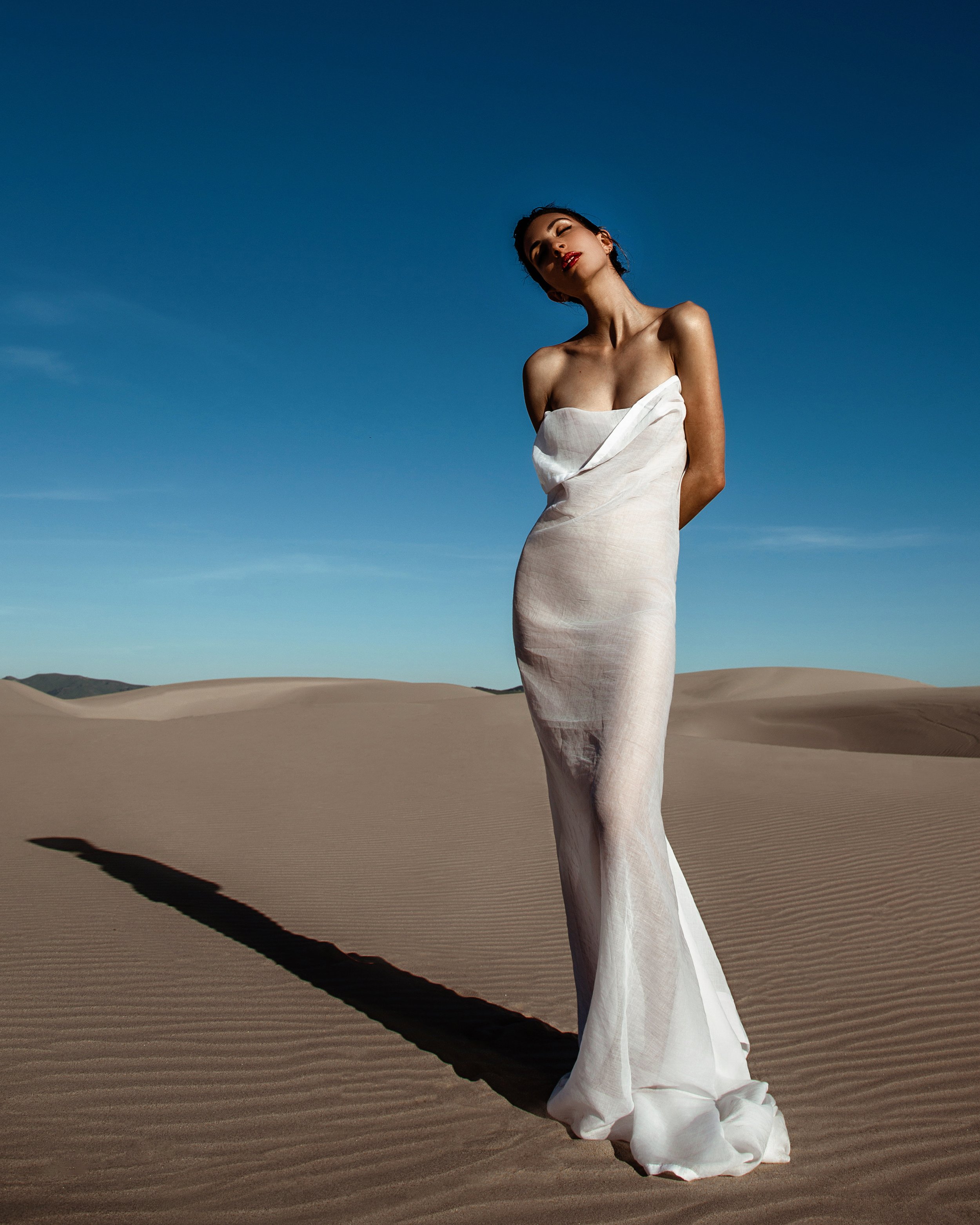 A woman in a white dress stands in a desert landscape with sand dunes, her eyes closed and head tilted back, casting a long shadow on the sand under a clear blue sky.