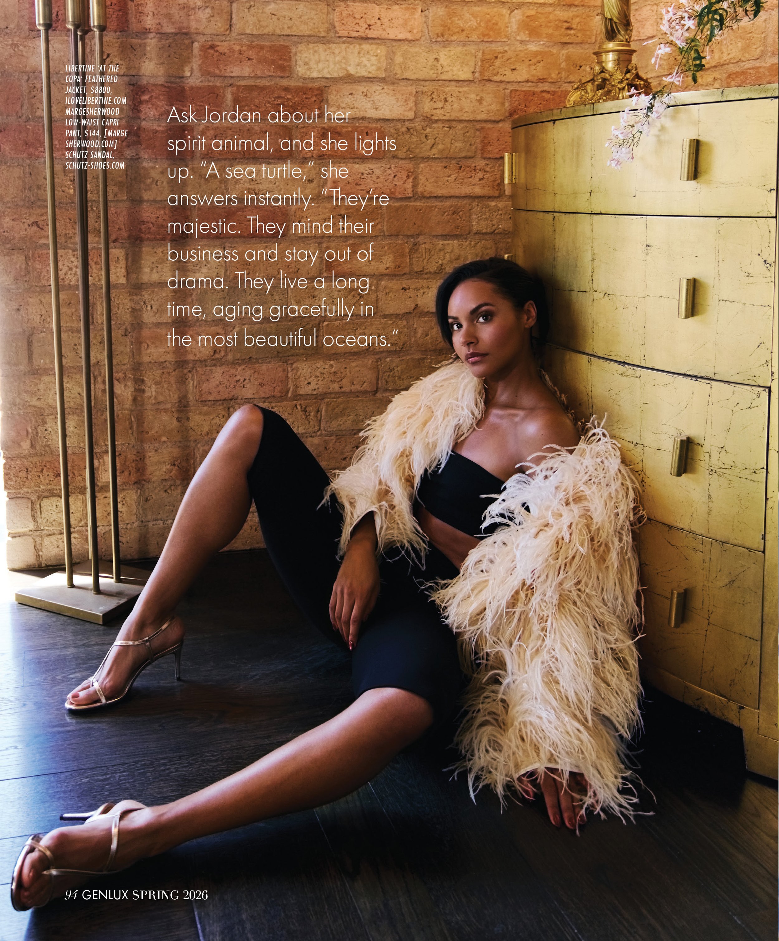 A woman, wearing high heels and a feathered coat, sitting on the floor with her back against a yellow dresser in a room with exposed brick walls. She is partially sitting on the floor with her legs extended and crossed at the ankles. The photo appear
