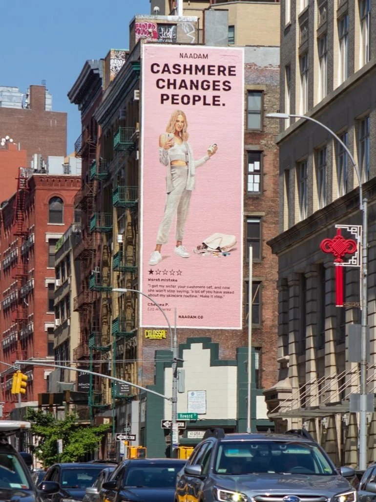 A large pink billboard on the side of a building features an image of a woman in a gray suit holding a drink and a phone, with a rollerboard at her feet. The billboard reads "NAADAM CASHMERE CHANGES PEOPLE." The surrounding cityscape includes brick a