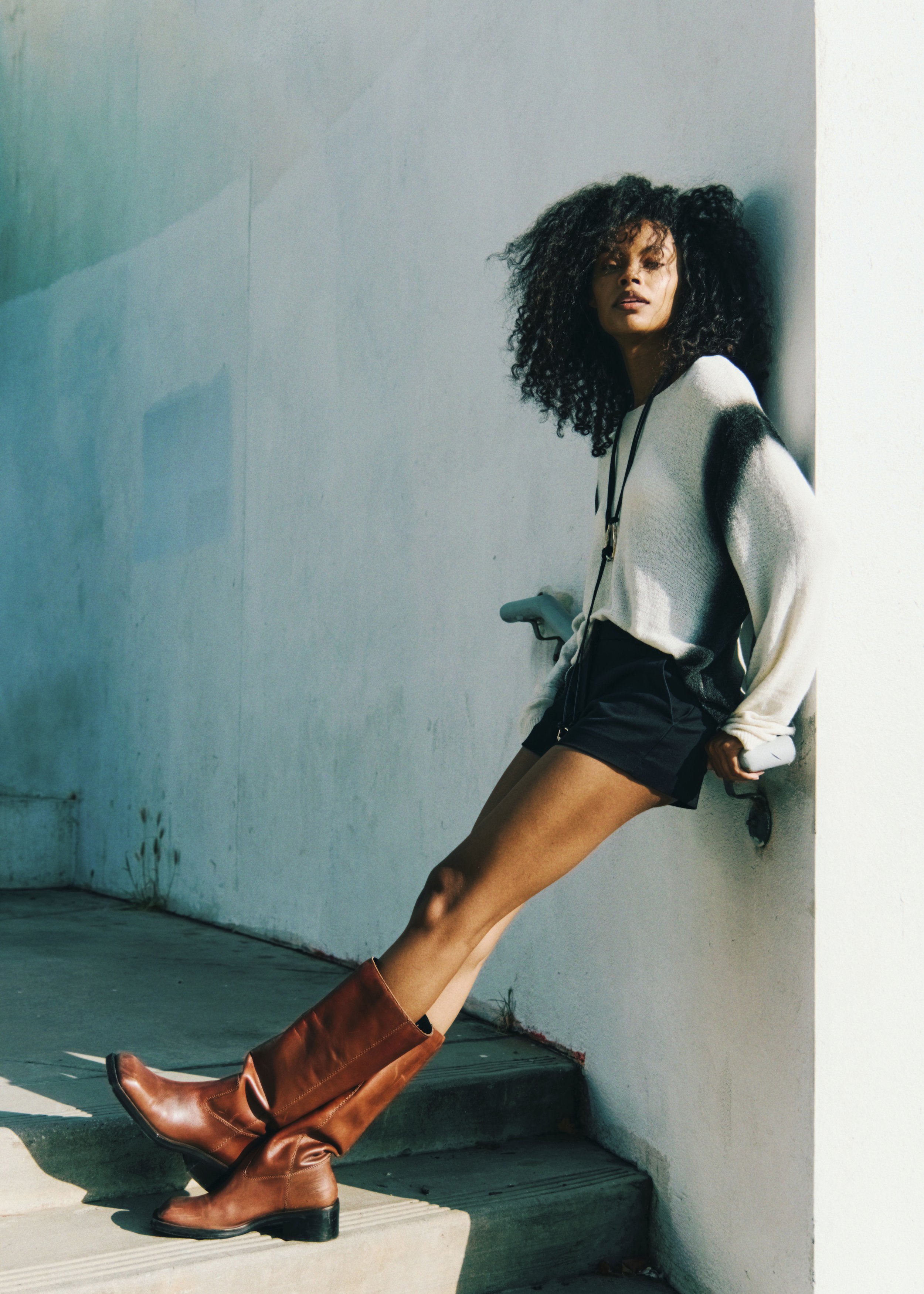 A woman with curly dark hair wearing a gray and black sweater, black shorts, and tall brown leather boots, standing against a plain white wall with sunlight casting shadows.