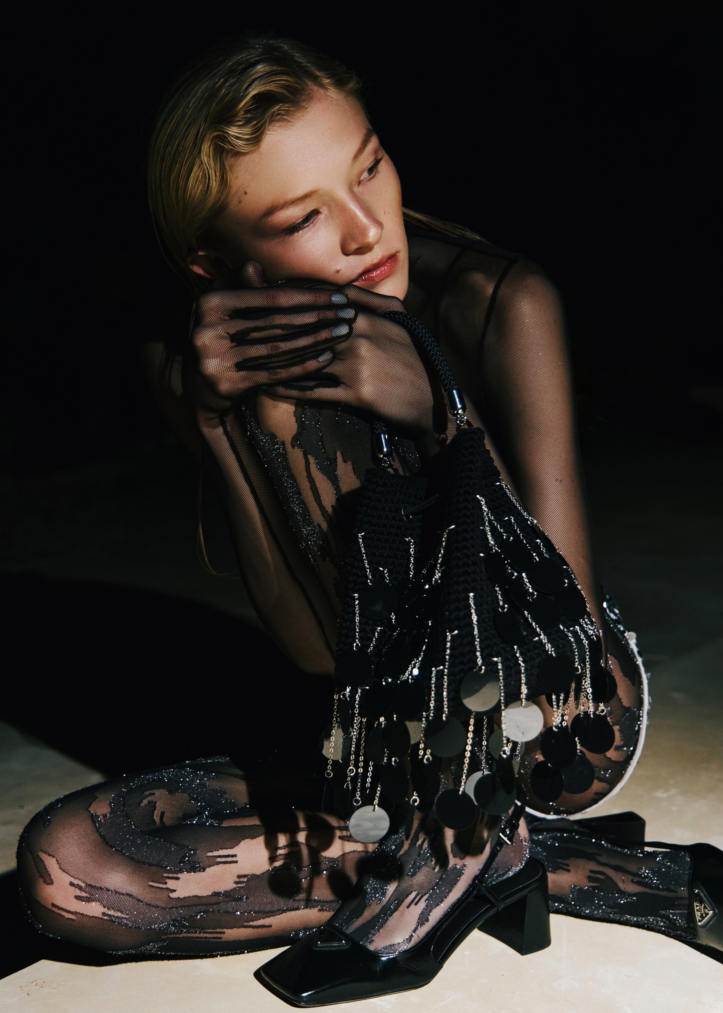 A young woman with blonde hair wearing a black, sheer, sequined dress with large circular embellishments, black gloves, and black high-heeled shoes, crouching on the ground against a black background.
