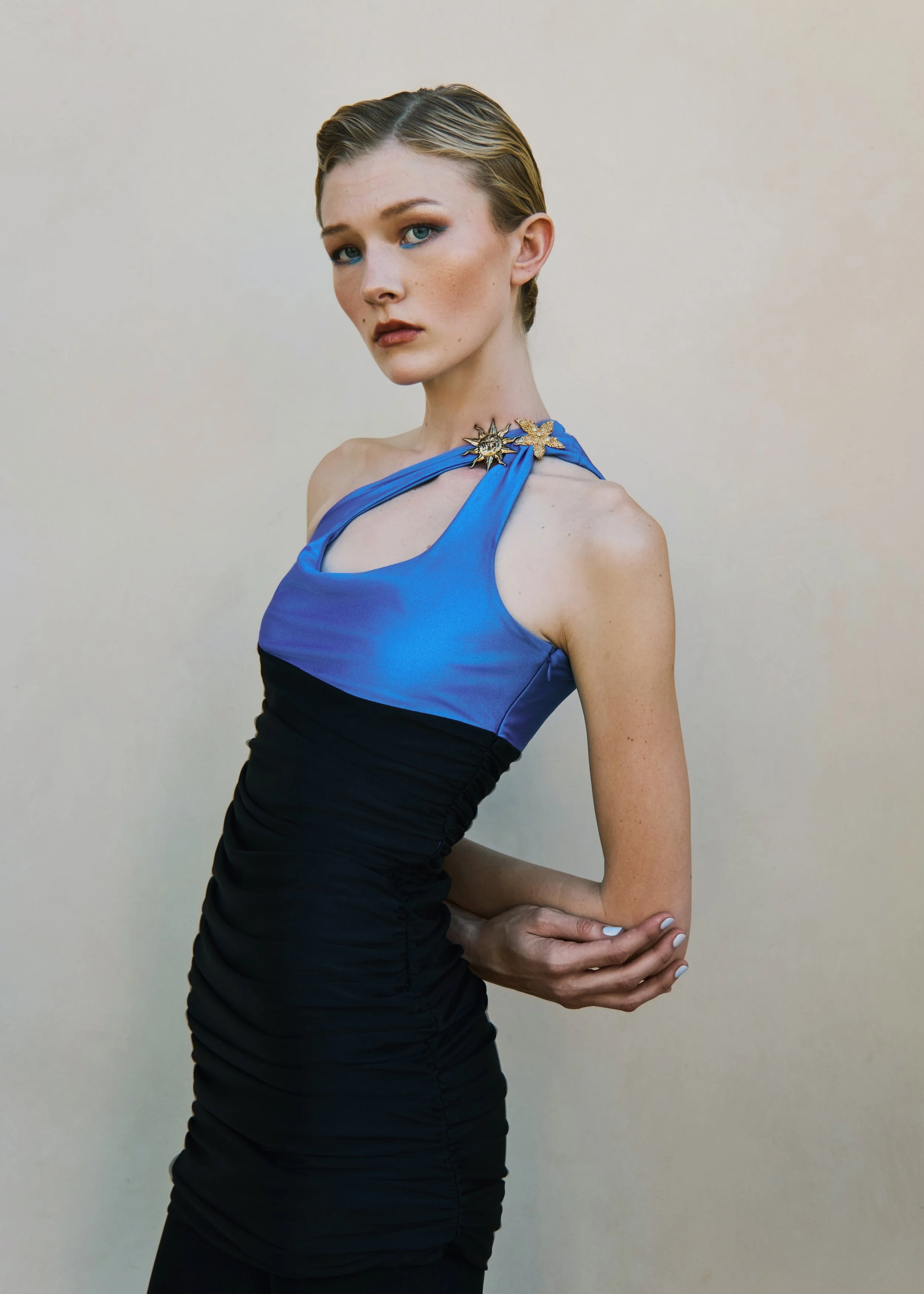 A woman with short, slicked-back blonde hair styled in waves, wearing a sleeveless, blue and black dress with decorative pins on the shoulder, standing against a plain, light-colored background.