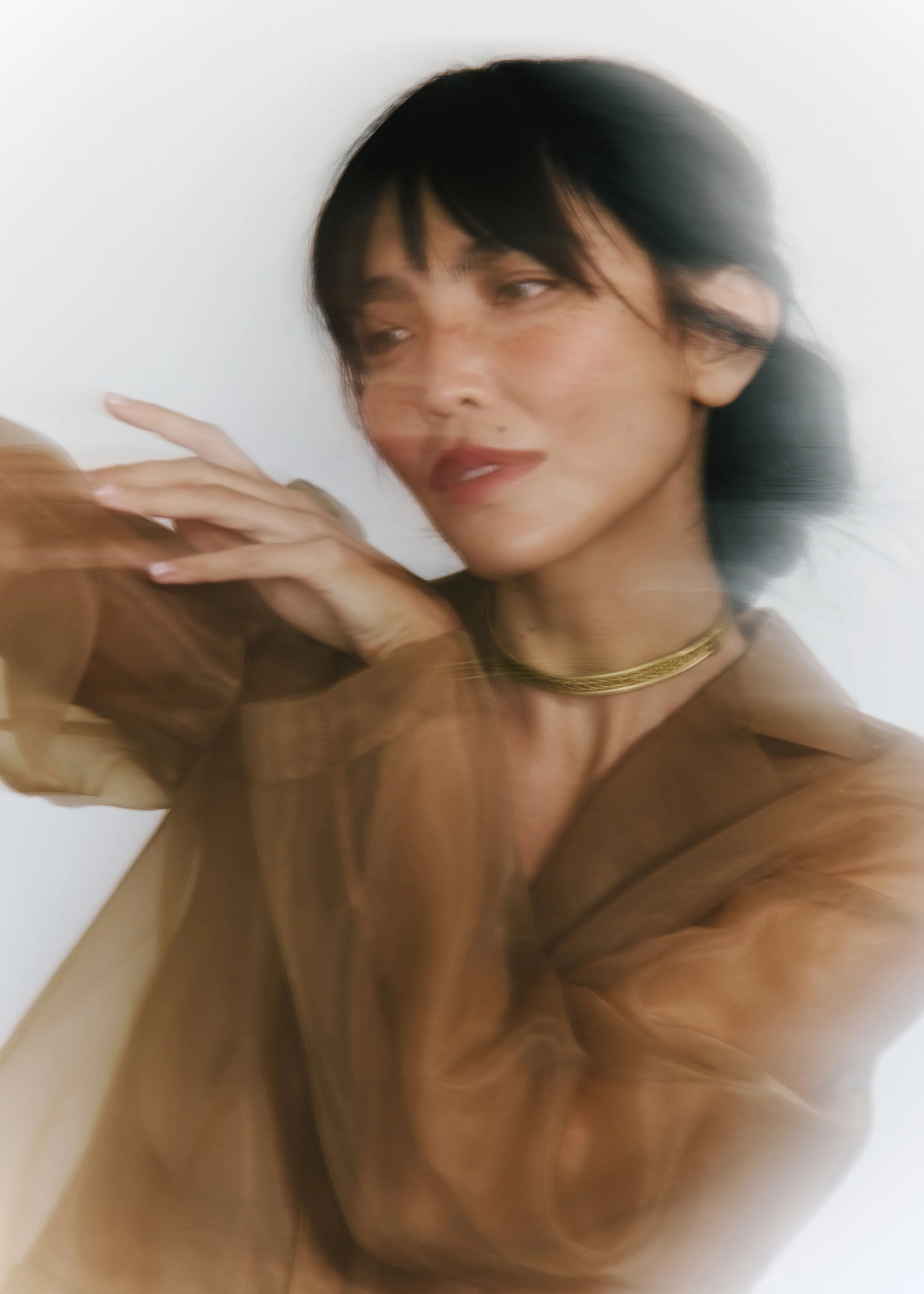 A woman with black hair and light skin taking a selfie, her head tilted slightly to the side, wearing a gold necklace, indoors with a plain white background.