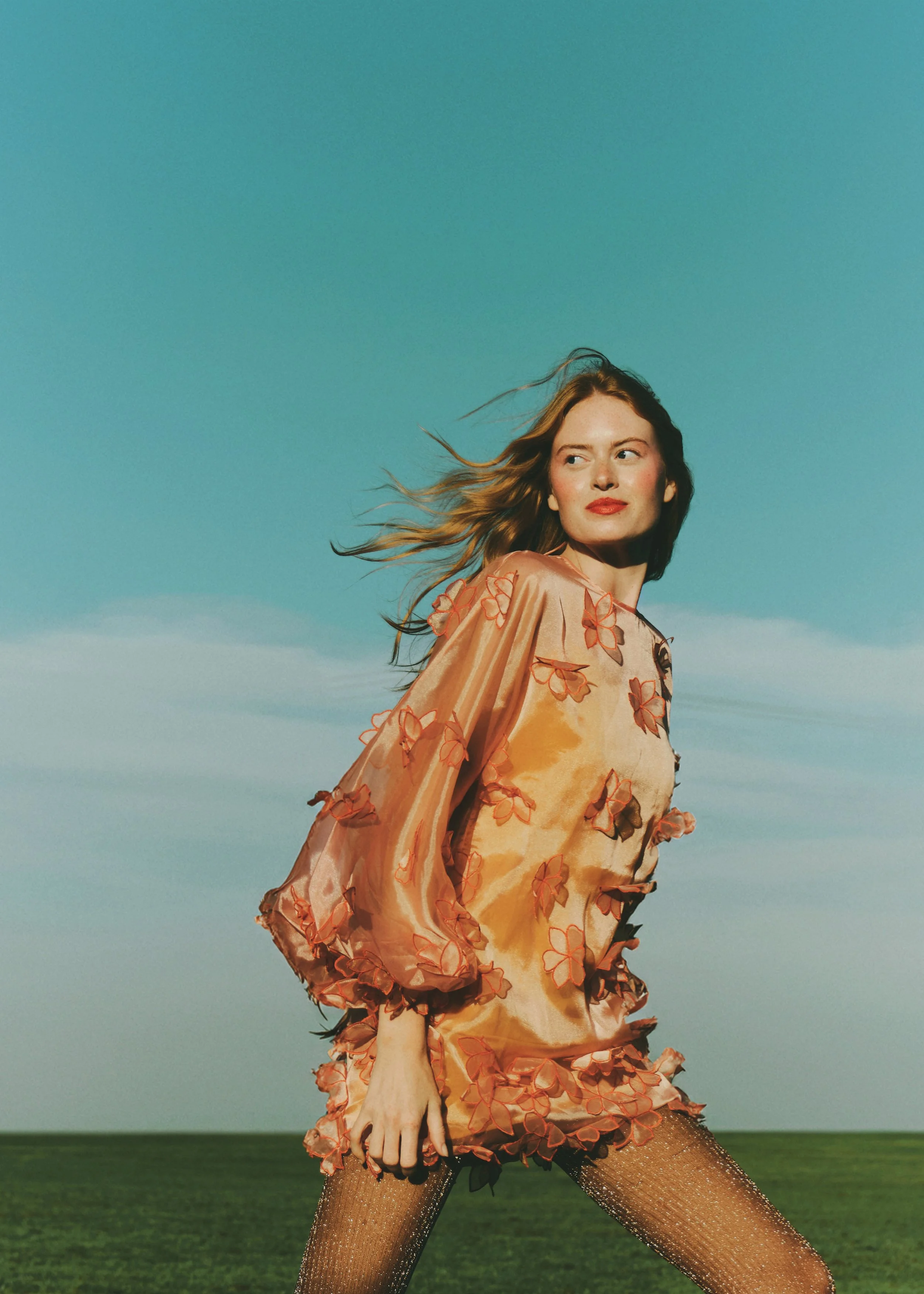 A woman with long hair standing outdoors on a grassy field under a clear blue sky, wearing a peach-colored dress with floral embroidery and glittery stockings.