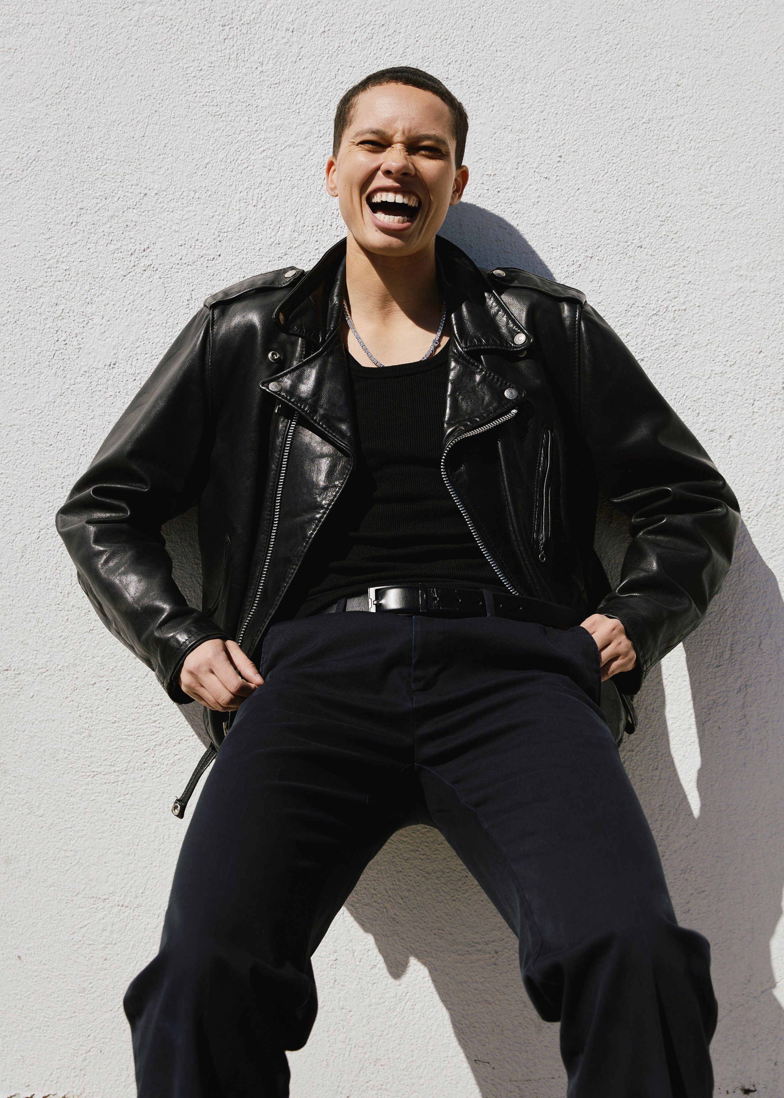 Young person wearing a black leather jacket, black shirt, and black pants, laughing or shouting, sitting against a plain white textured wall, with legs spread apart and hands resting on thighs.
