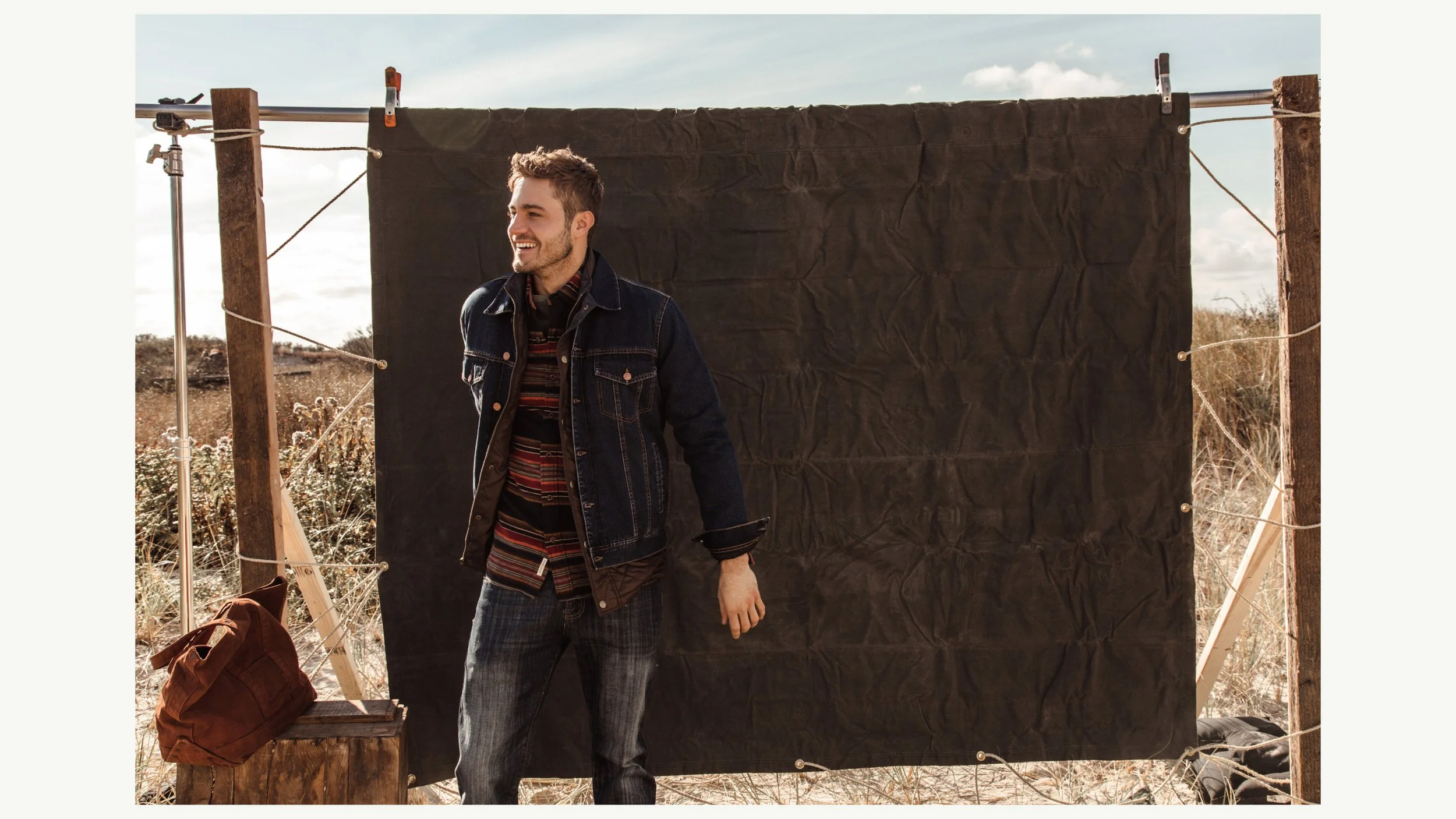 A smiling man stands outdoors in front of a black fabric backdrop propped up on wooden and metal supports, with dry grass and plants in the background.