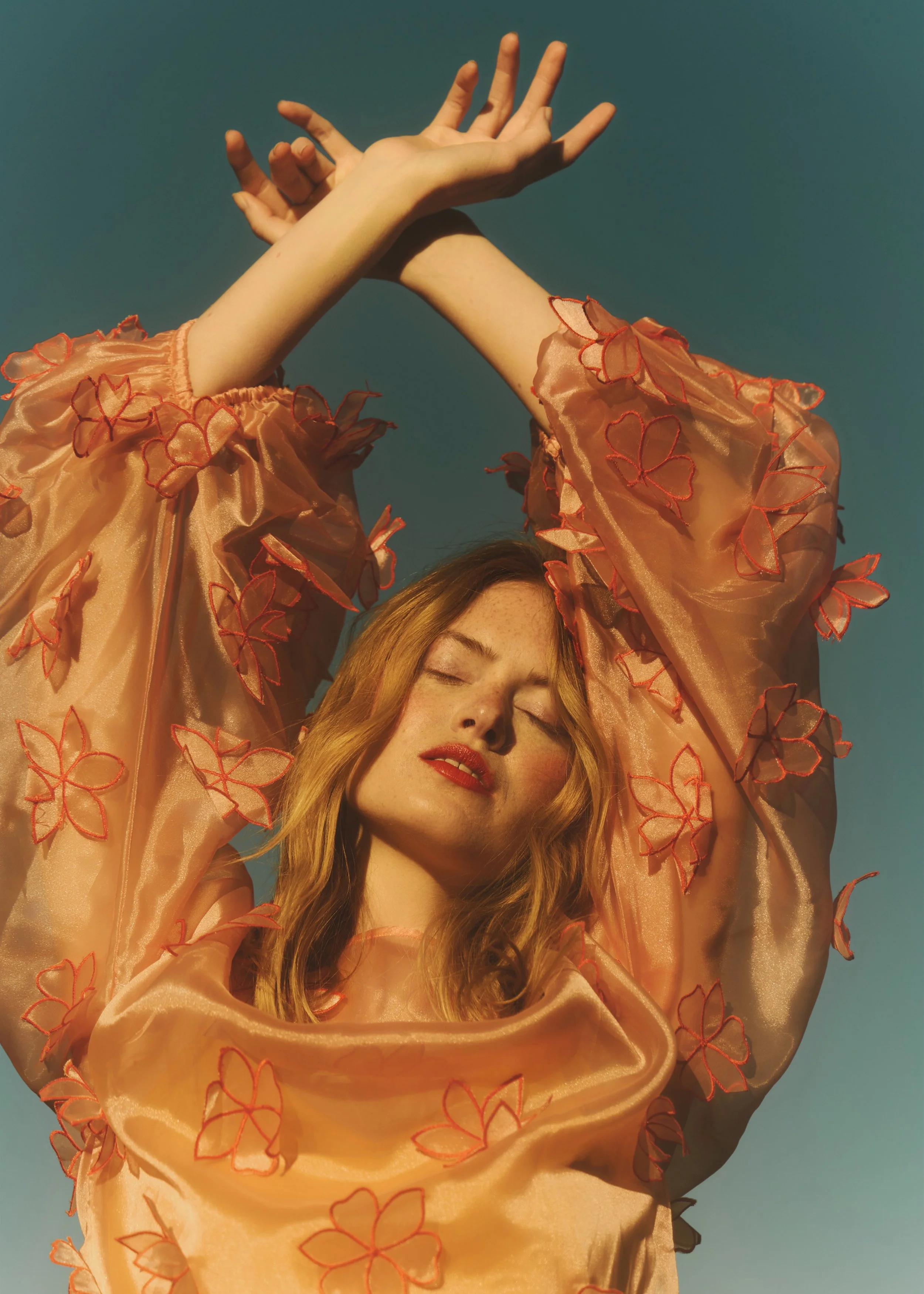 A woman with blonde hair and red lipstick is raising her arms above her head, wearing a peach-colored, sheer dress with red embroidered flower patterns, against a clear blue sky.