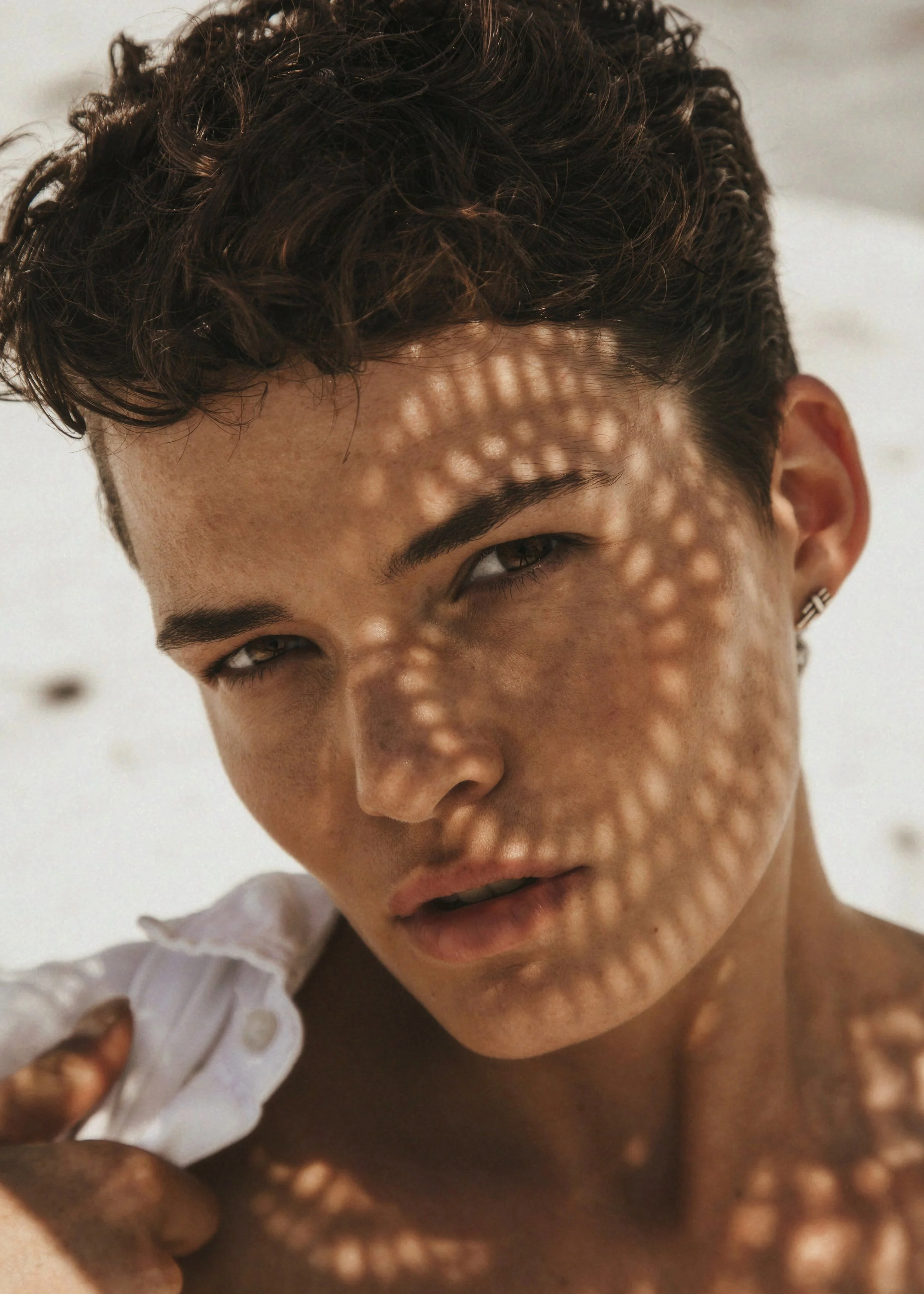 Close-up of a person's face with textured skin, dark curly hair, and a single hoop earring, partially covering one eye while holding a white cloth to the face.
