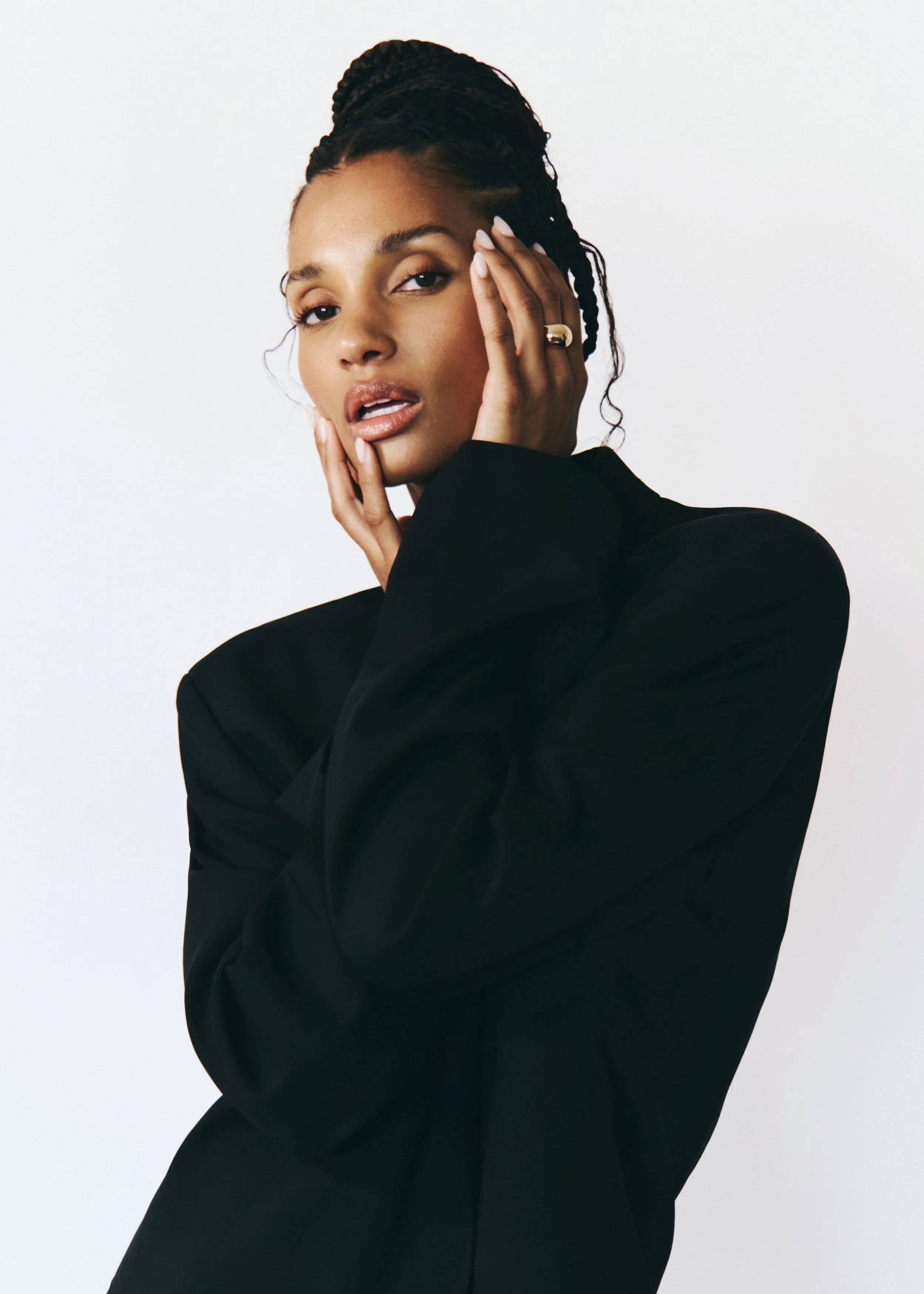 A woman with braided hair, wearing a black outfit, posing with one hand on her face, looking at the camera against a plain white background.