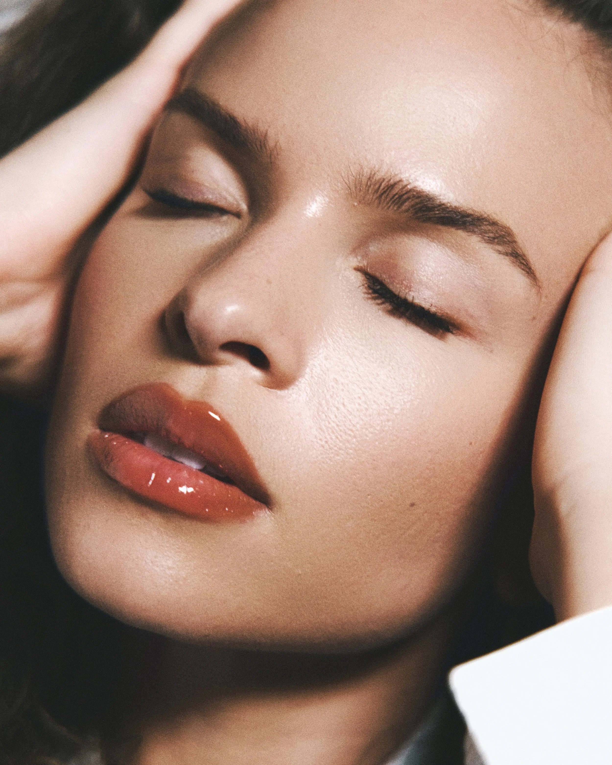 Close-up of a woman's face with closed eyes, natural makeup, and slightly parted lips. She is touching her head with her hands.