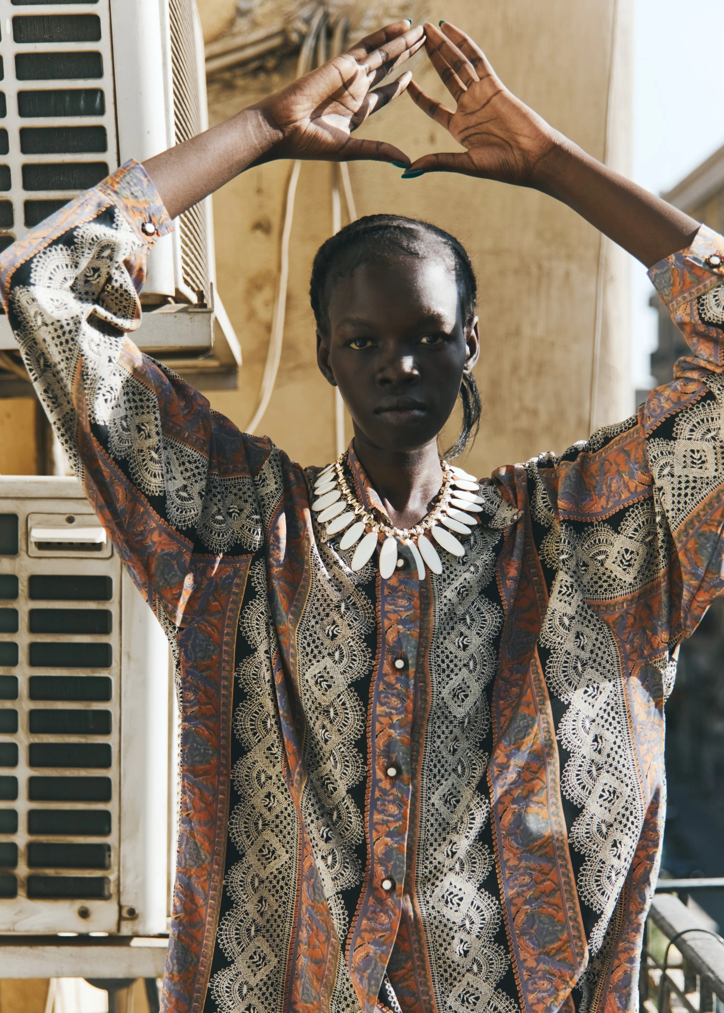 A woman with dark skin and braided hair making a heart shape with her hands, wearing a patterned dress and a large shell necklace, standing outdoors near a building wall.