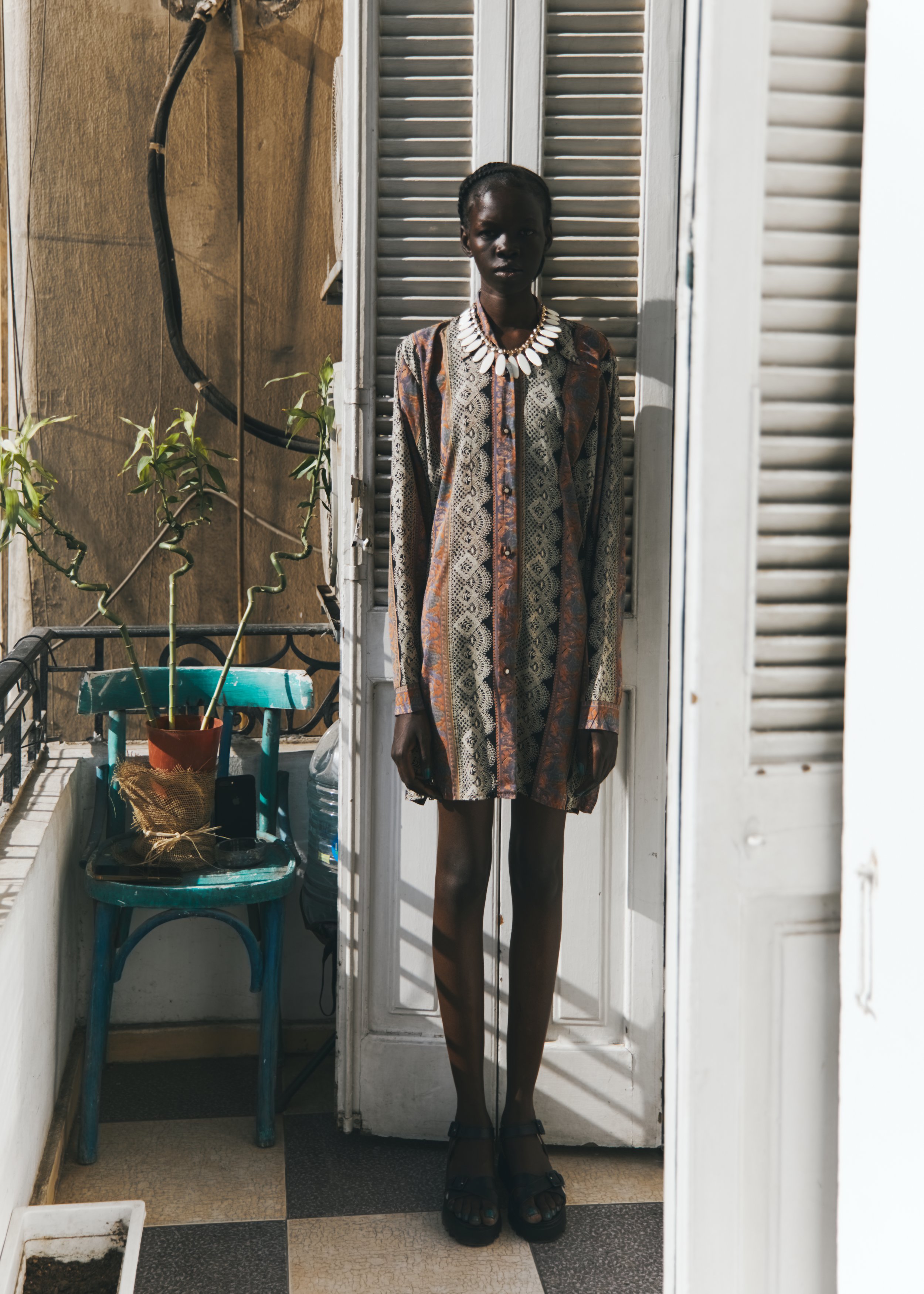 A person with dark skin standing indoors against a closed louvered door, wearing a patterned dress, black sandals, and a white necklace. To the left, there is a small table with a potted plant and some objects.