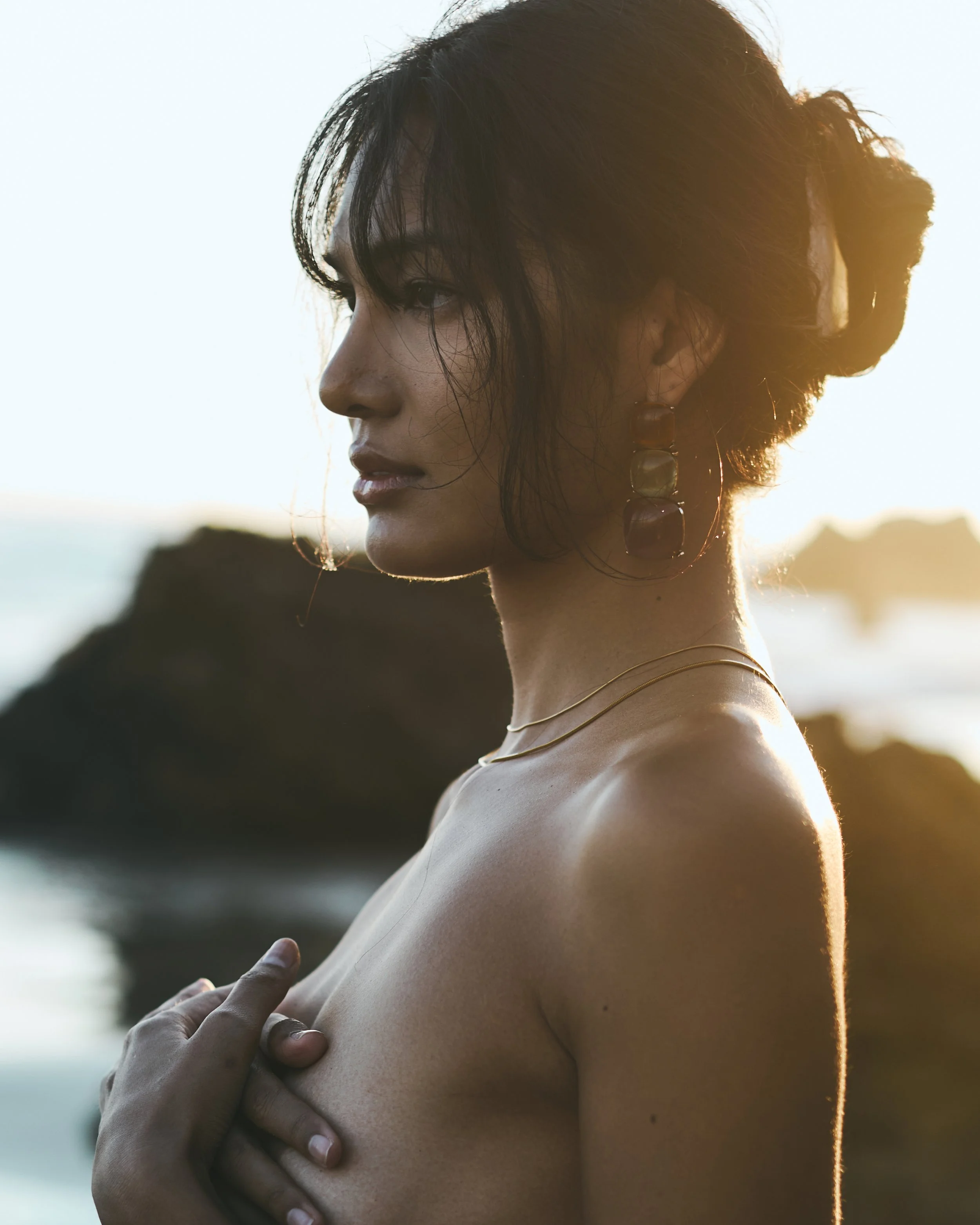 Profile of a woman with short dark hair, wearing large earrings and a delicate necklace, standing outdoors with a sunset in the background.