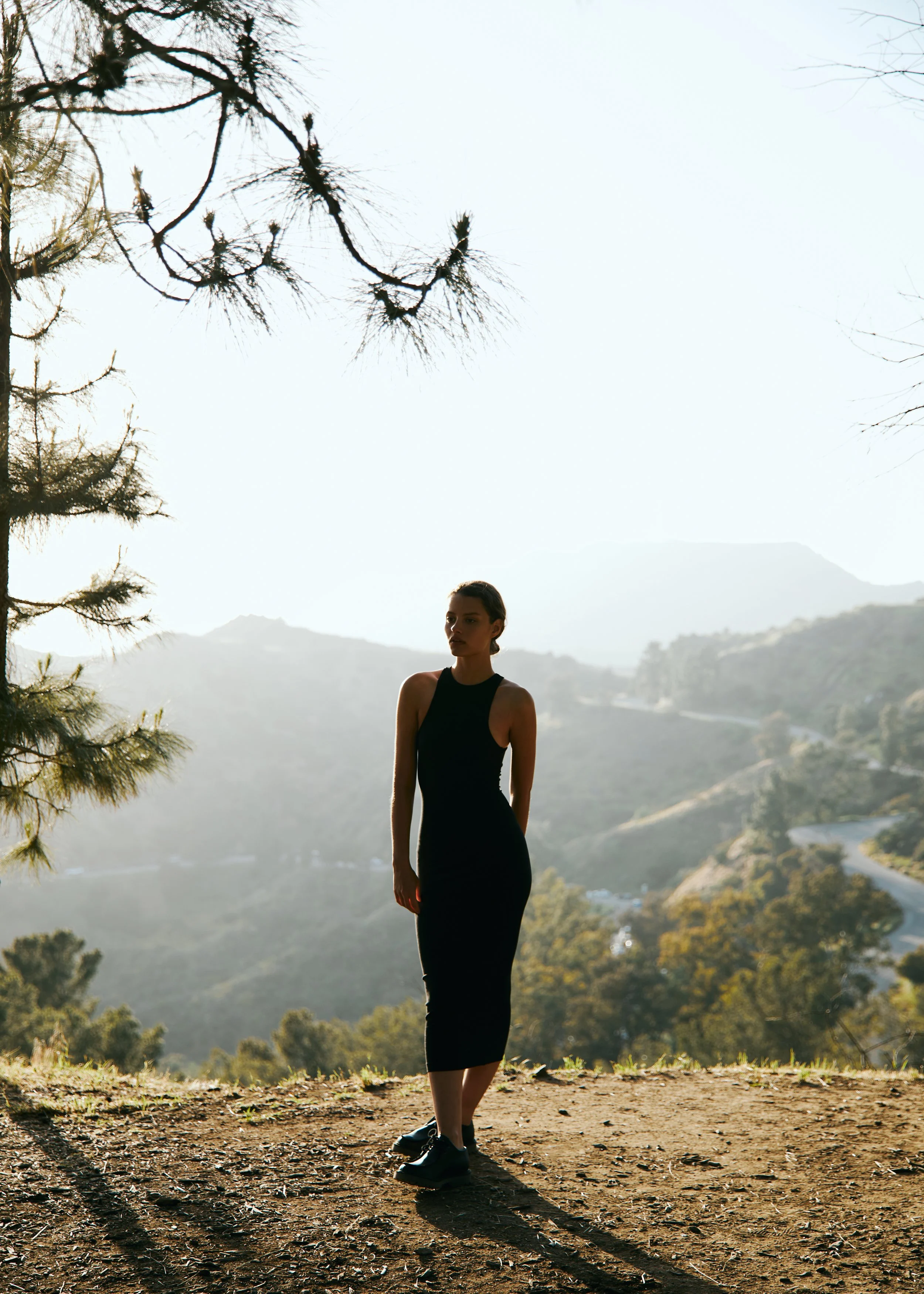 A woman standing outdoors on a dirt path with mountains and trees in the background, wearing a black sleeveless dress and sneakers, with sunlight casting her shadow on the ground.