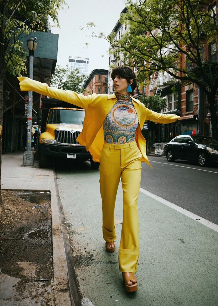 A woman in a yellow outfit walking on a city sidewalk with her arms outstretched, surrounded by urban buildings, trees, and parked cars.