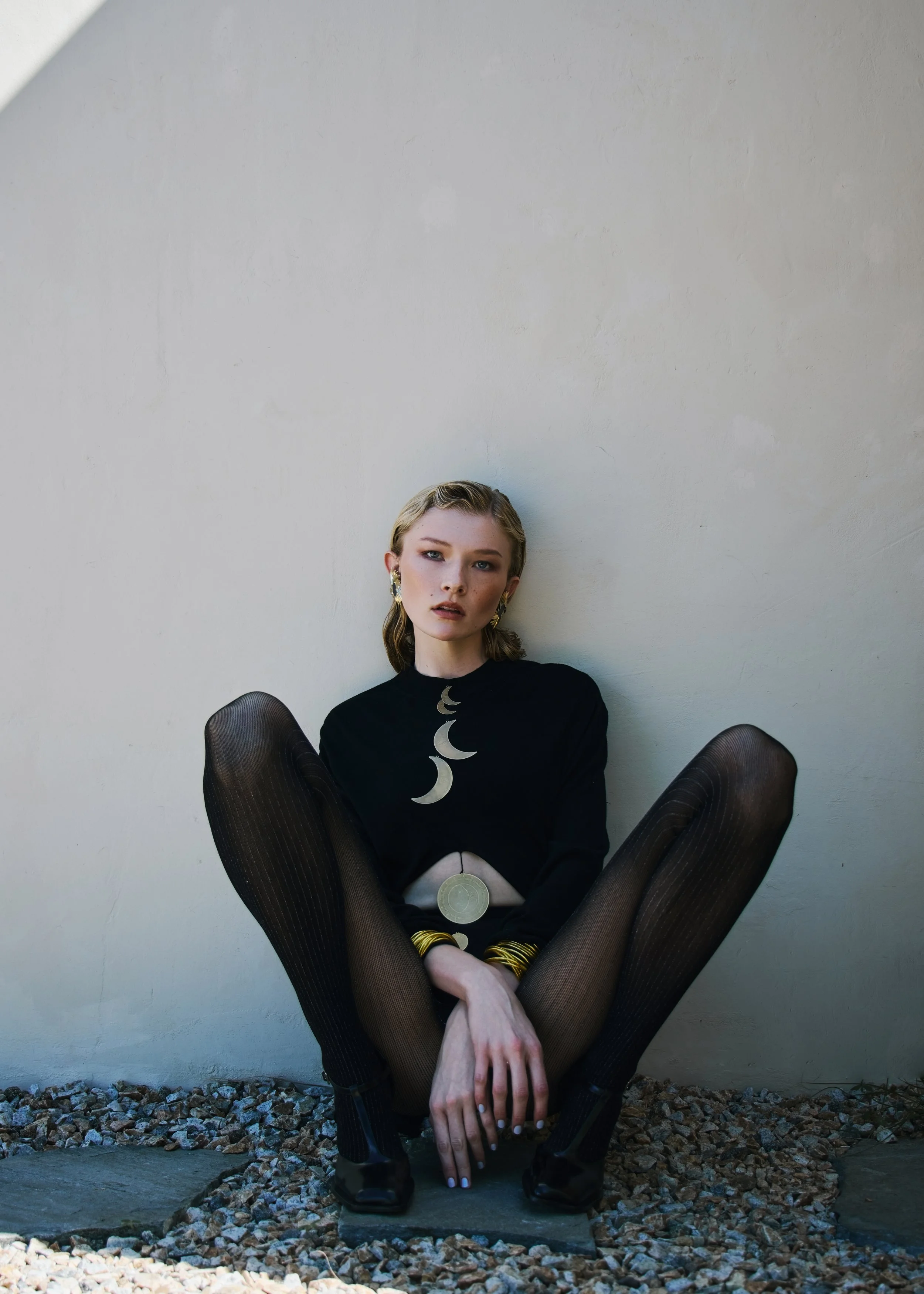 Young woman sitting cross-legged on the ground, leaning against a white wall, wearing a black top with moon phases design, black fishnet tights, black shoes, gold jewelry, and earrings, with her hair in waves and makeup.