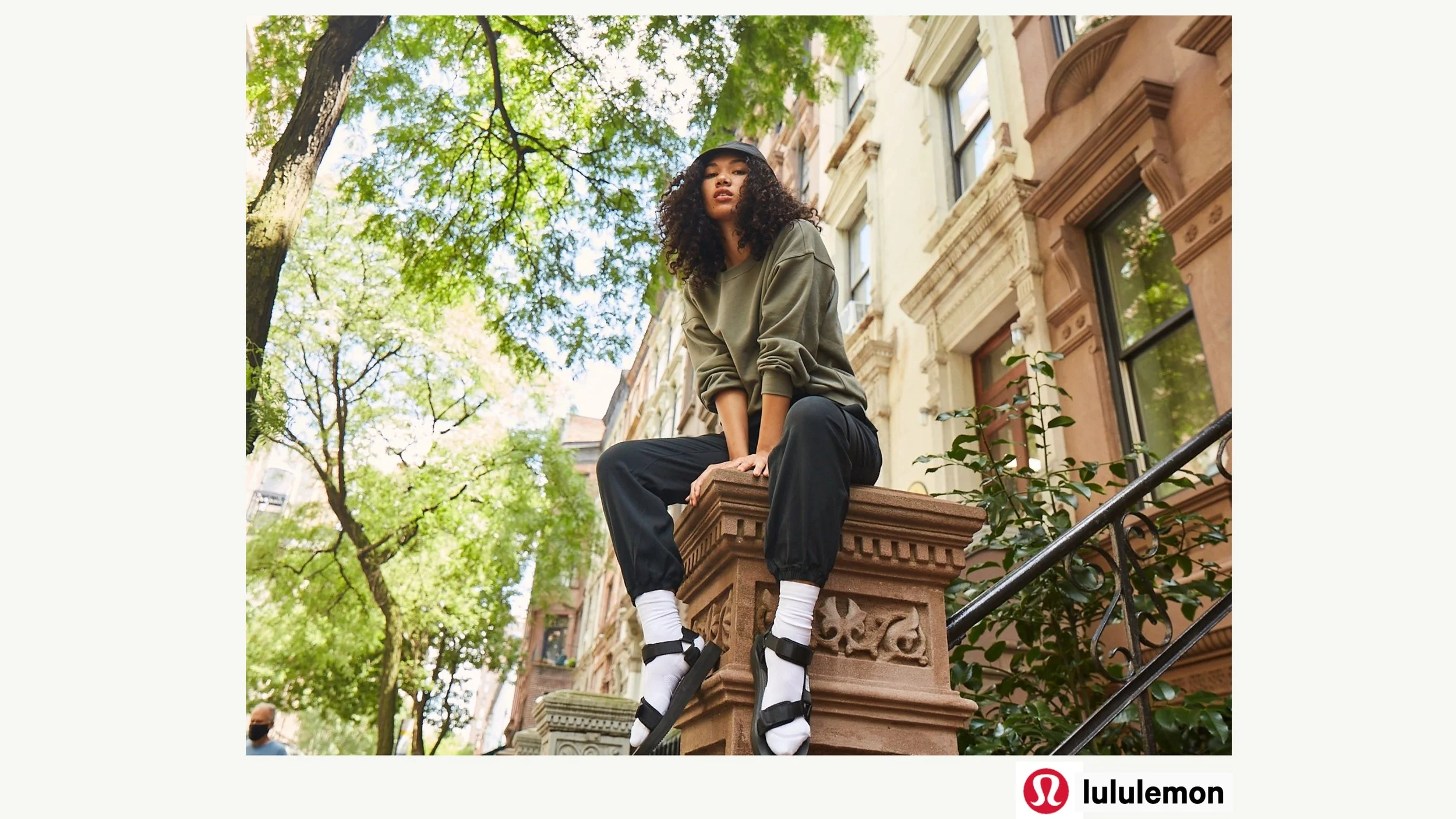 A young woman with curly hair, wearing a black cap, green sweatshirt, black pants, and white socks with black sandals, sitting on the edge of a brownstone building's stoop in a city neighborhood. She is looking at the camera with a neutral expression
