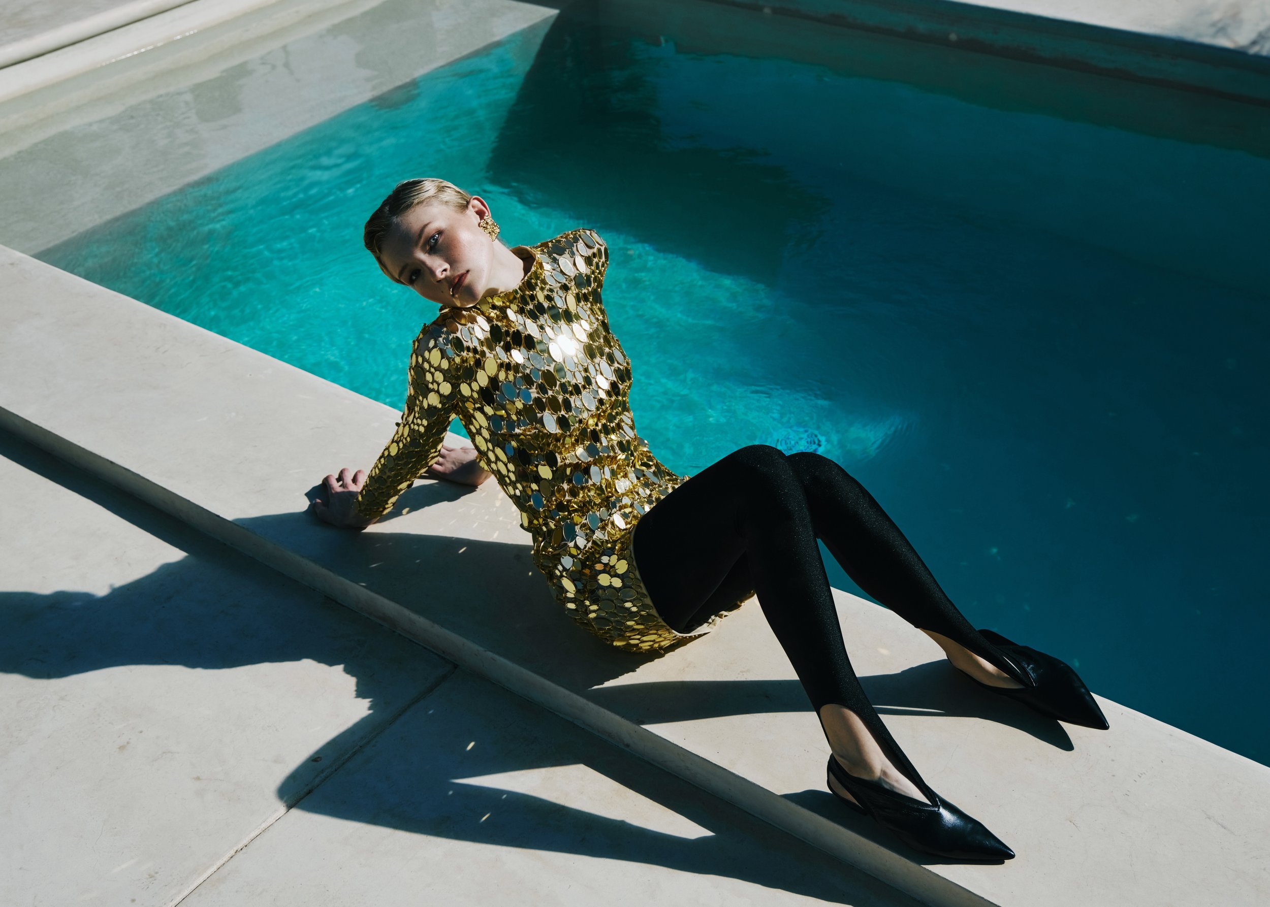 A woman in a shiny gold and black dress with black high heels, sitting on the edge of a swimming pool, looking at the camera.