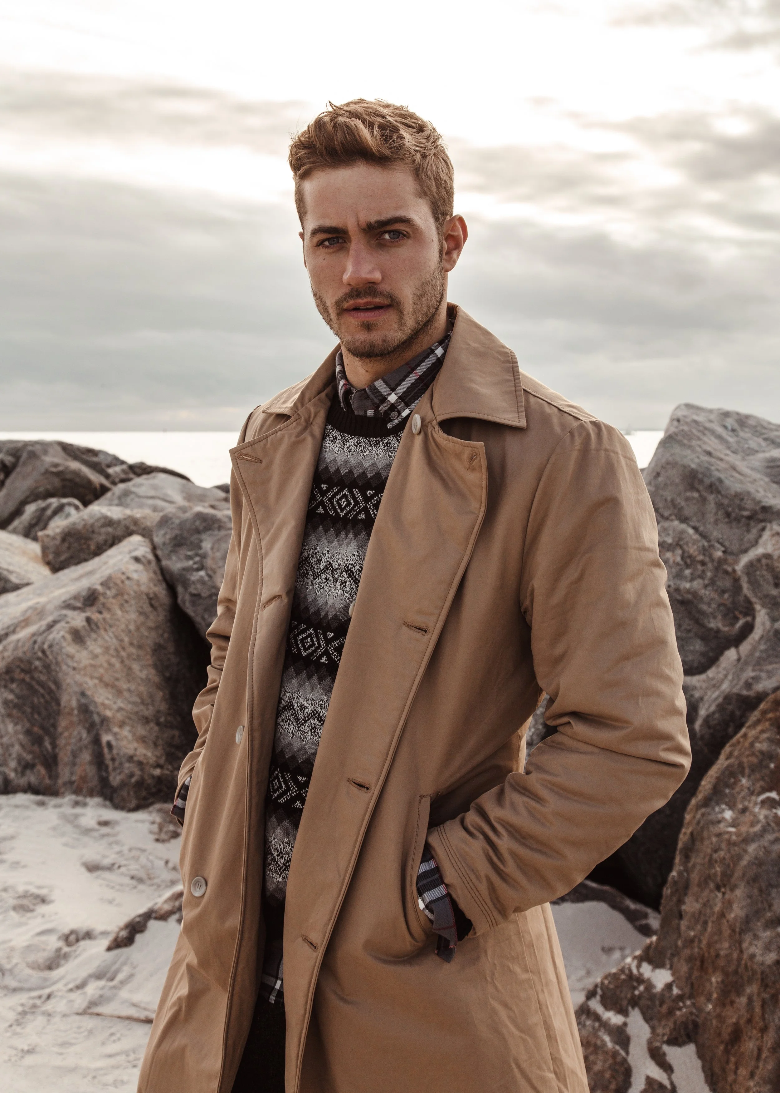 A young man with short brown hair and a slight beard standing outdoors near rocks, looking into the camera with a serious expression. He is wearing a tan trench coat, a hooded sweater, a plaid shirt, and a patterned sweater underneath. Overcast cloud