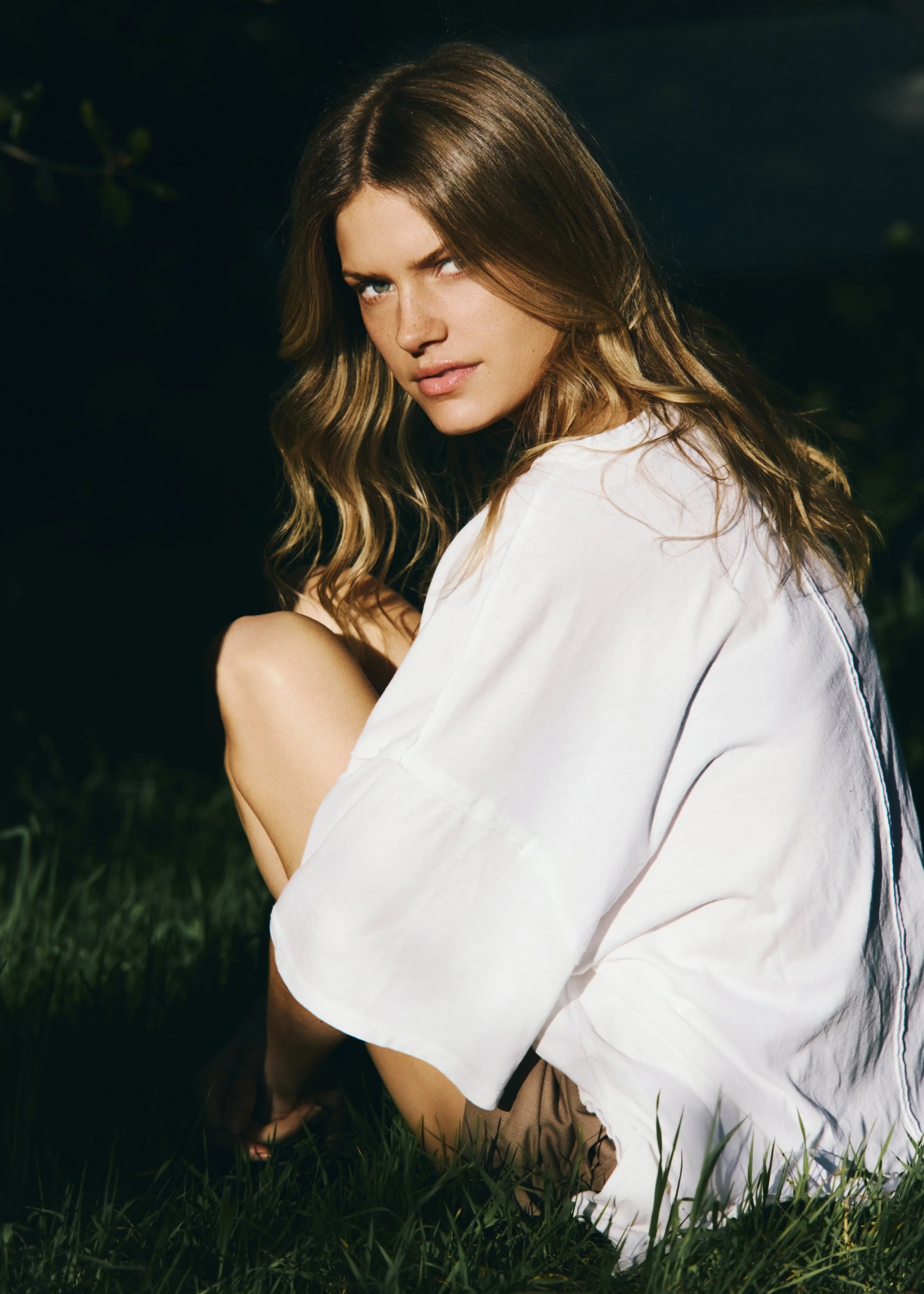 A young woman with long wavy light brown hair, green eyes, and light skin, sitting outdoors in grass with a dark background, looking at the camera with a slight smile, wearing a white shirt.