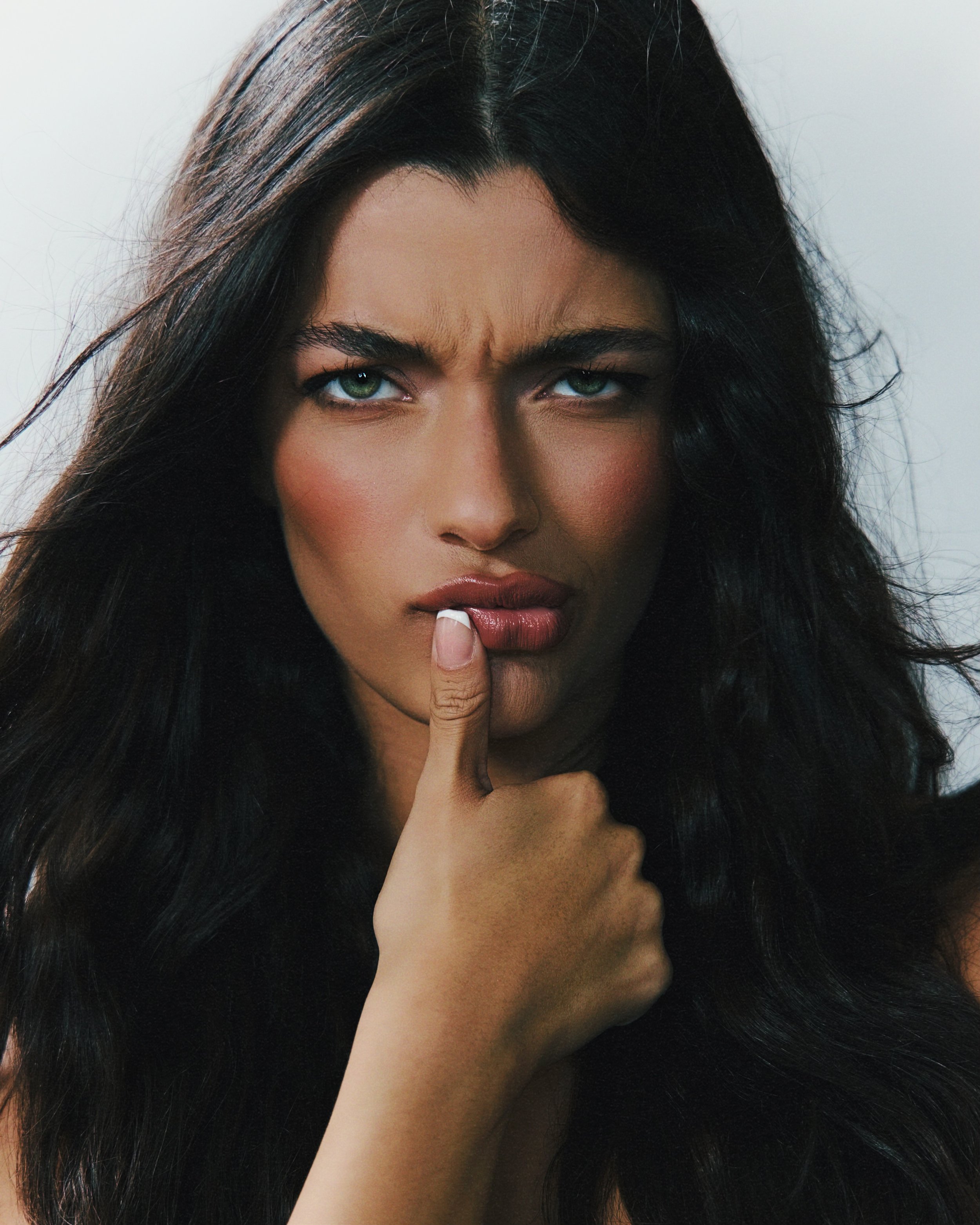 Close-up of woman with long dark hair, green eyes, and finger pressed against lips in gesture of silence.