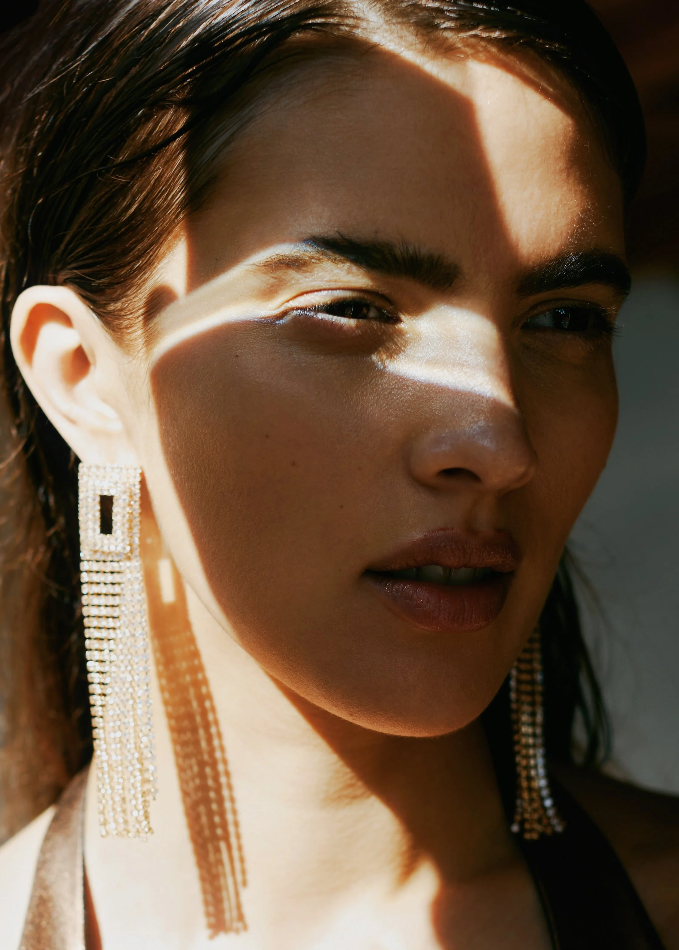 Close-up portrait of a woman with dark hair wearing large, sparkling earrings, with sunlight creating shadows on her face.