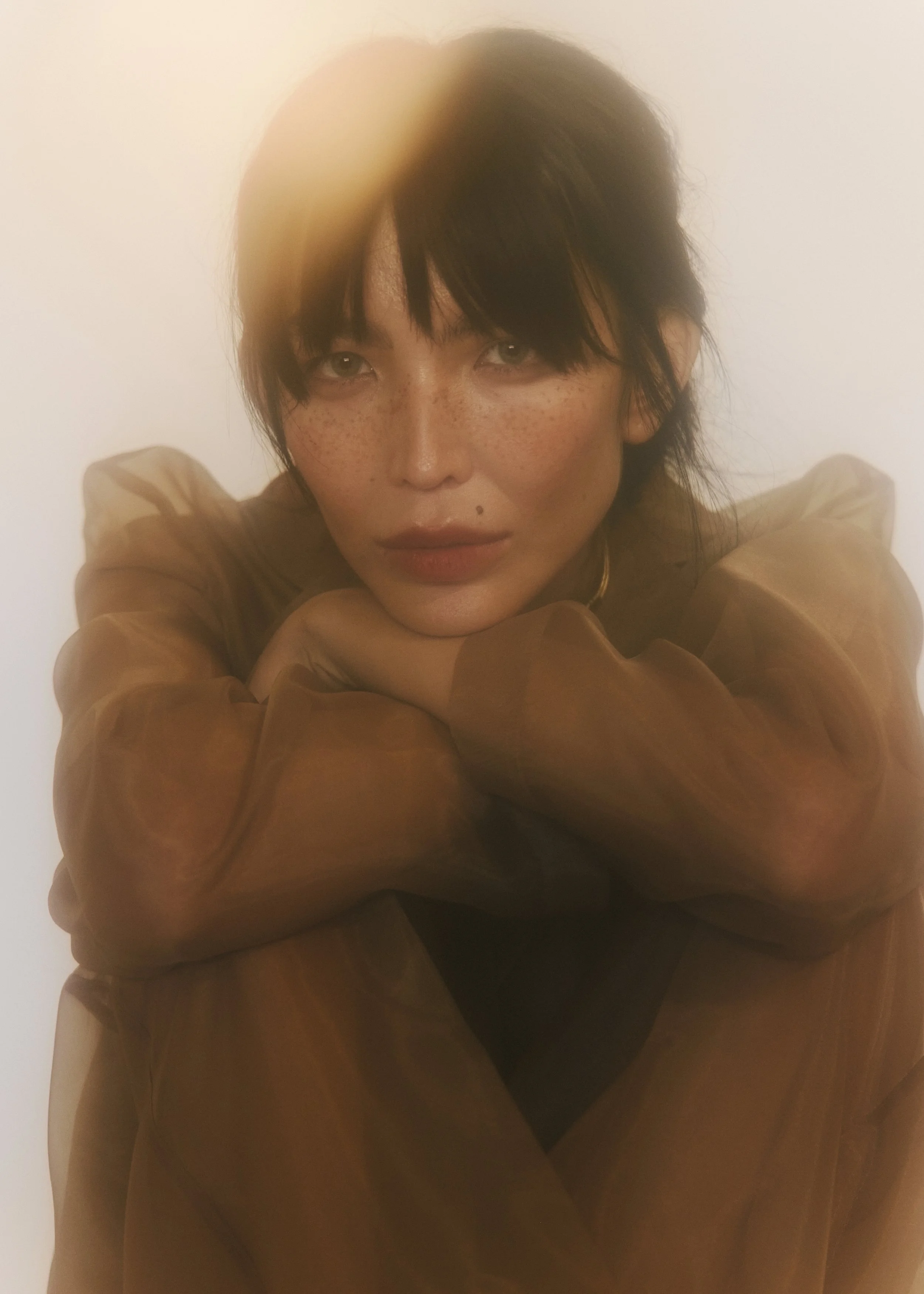 A person with dark, wavy hair and light skin, wearing a brown jacket, leaning forward with their head resting on their crossed arms, gazing into the camera.