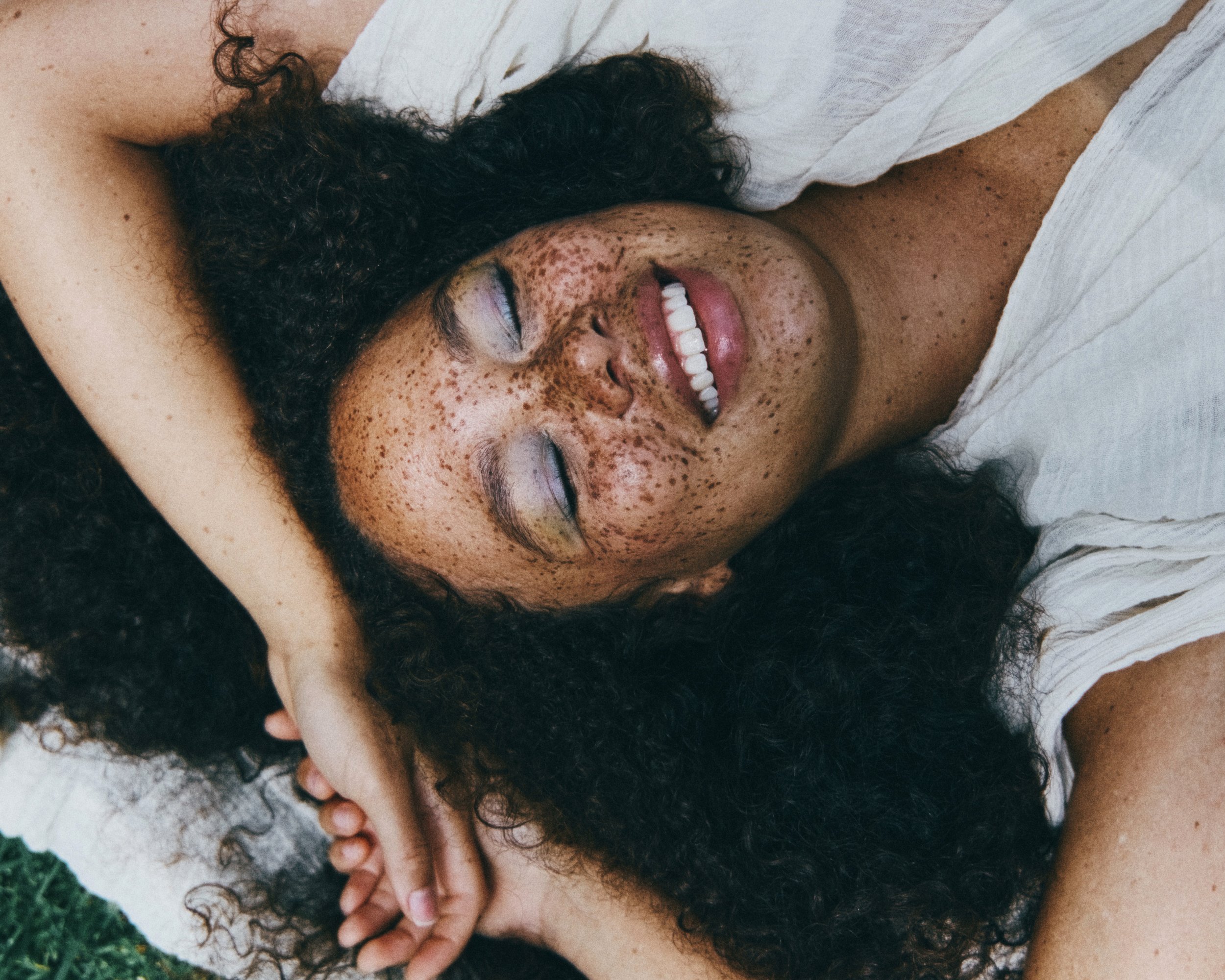 A woman with curly dark hair and freckles lying down on a white surface, smiling with her eyes closed, wearing a white shirt.