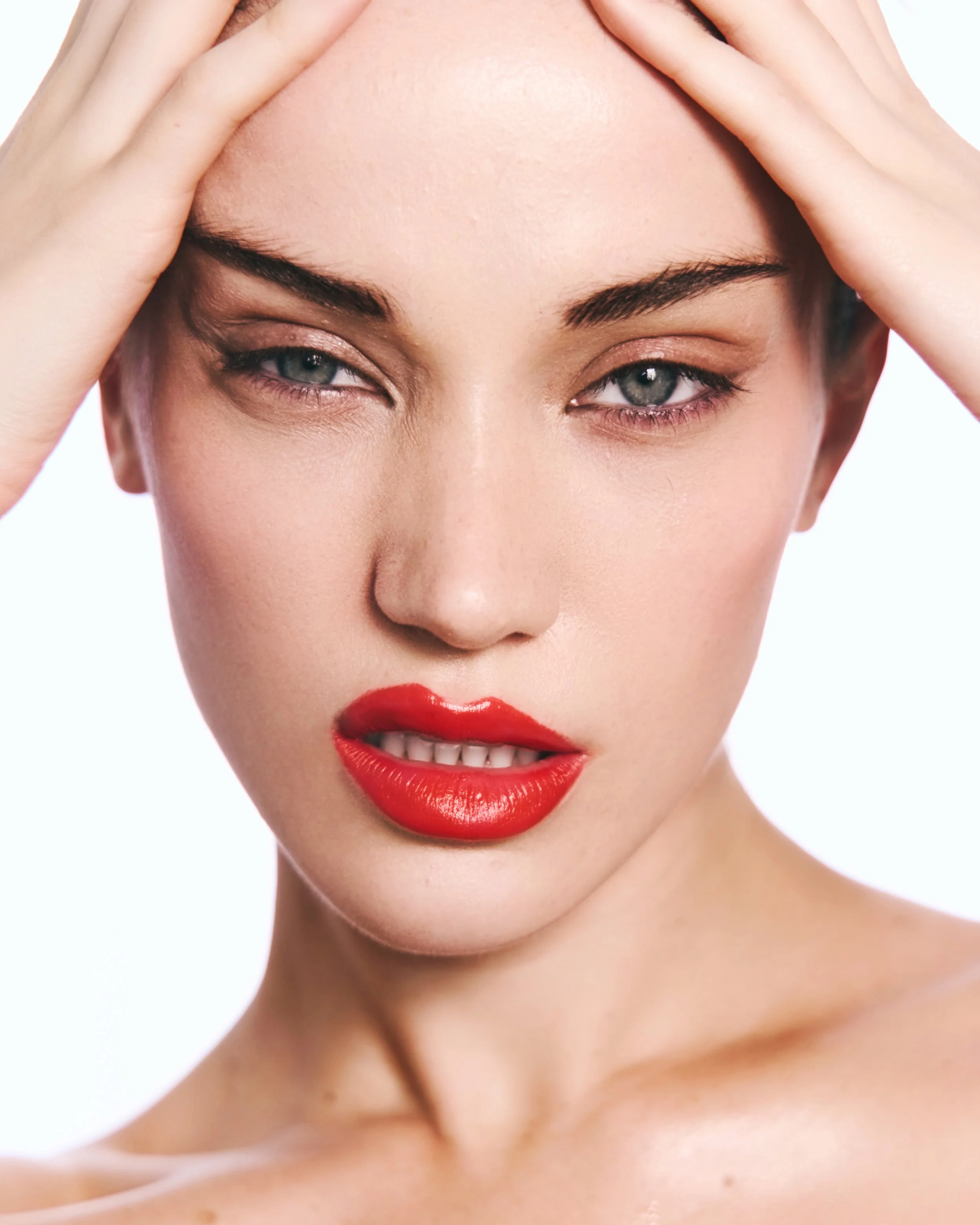 Close-up of a woman with fair skin, blue eyes, red lipstick, and lightly applied eye makeup. She is holding her head with both hands, fingers on her forehead, and looking slightly to the side.