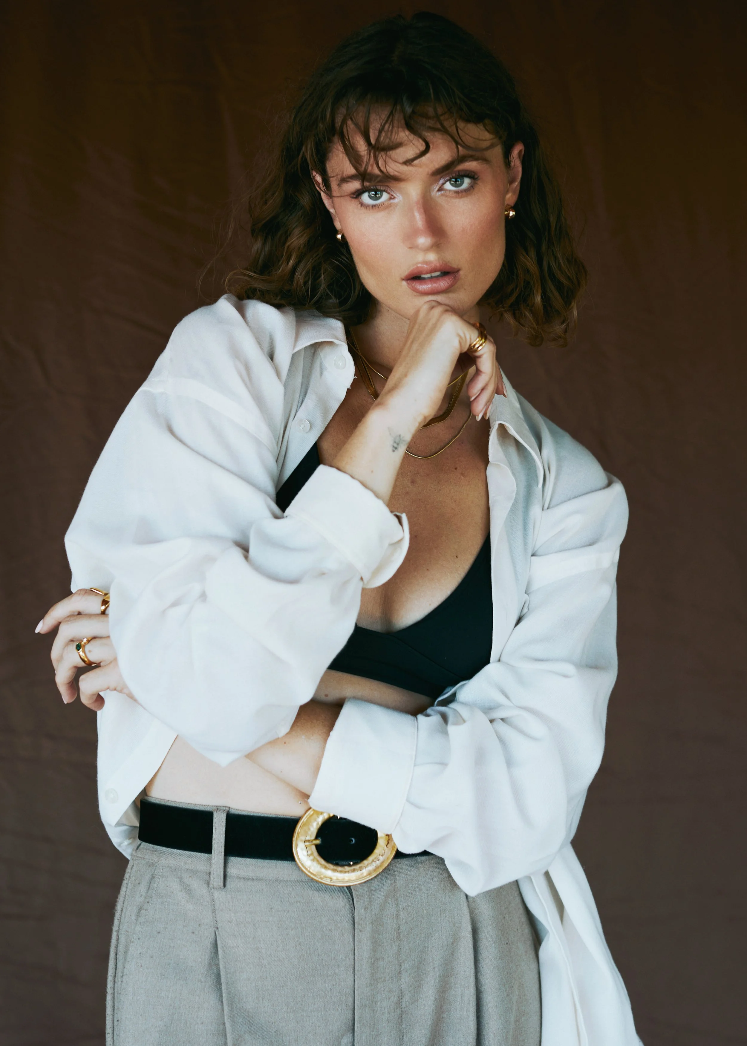 A woman with wavy brown hair, blue eyes, and fair skin is wearing a partially open white shirt, black top, grey high-waisted trousers, and a large black and gold belt. She is accessorized with gold jewelry, including earrings, rings, necklaces, and a