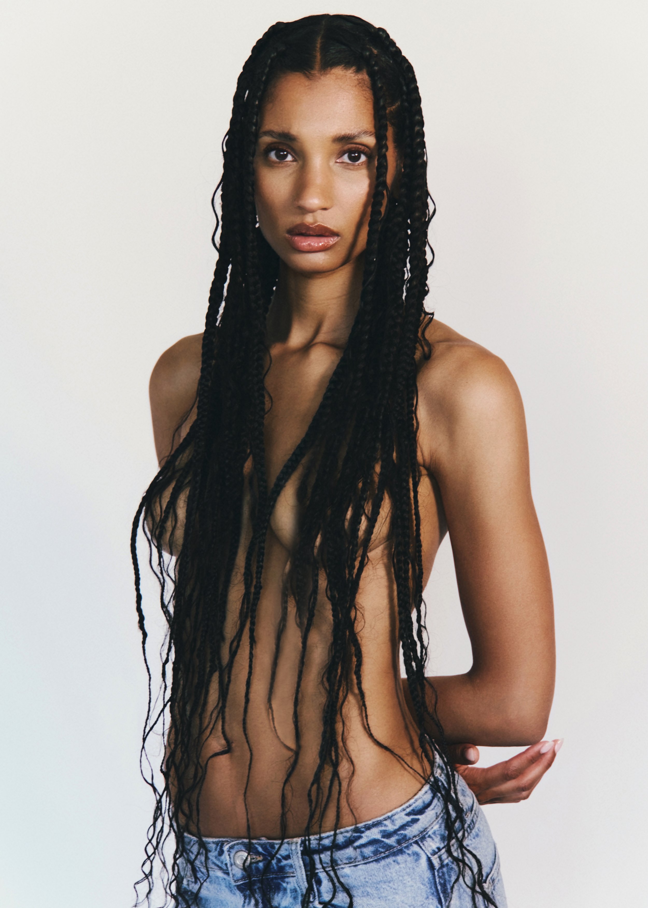 A woman with long braided hair and bare shoulders wearing high-waisted denim jeans against a plain white background.