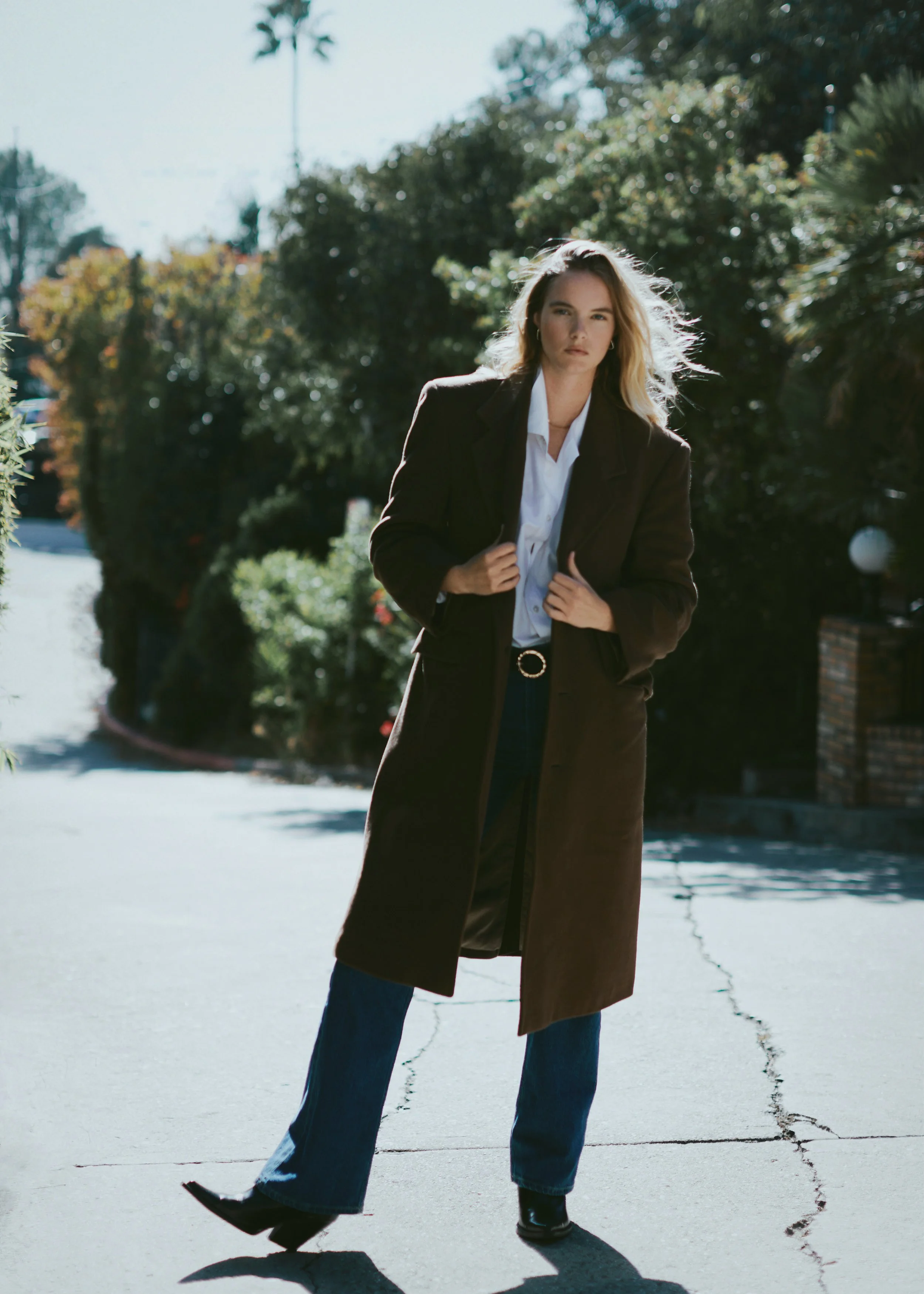 A woman with blonde hair, wearing a long brown coat, white blouse, blue jeans, and black shoes, walking on a cracked sidewalk outdoors with green bushes and palm trees in the background.