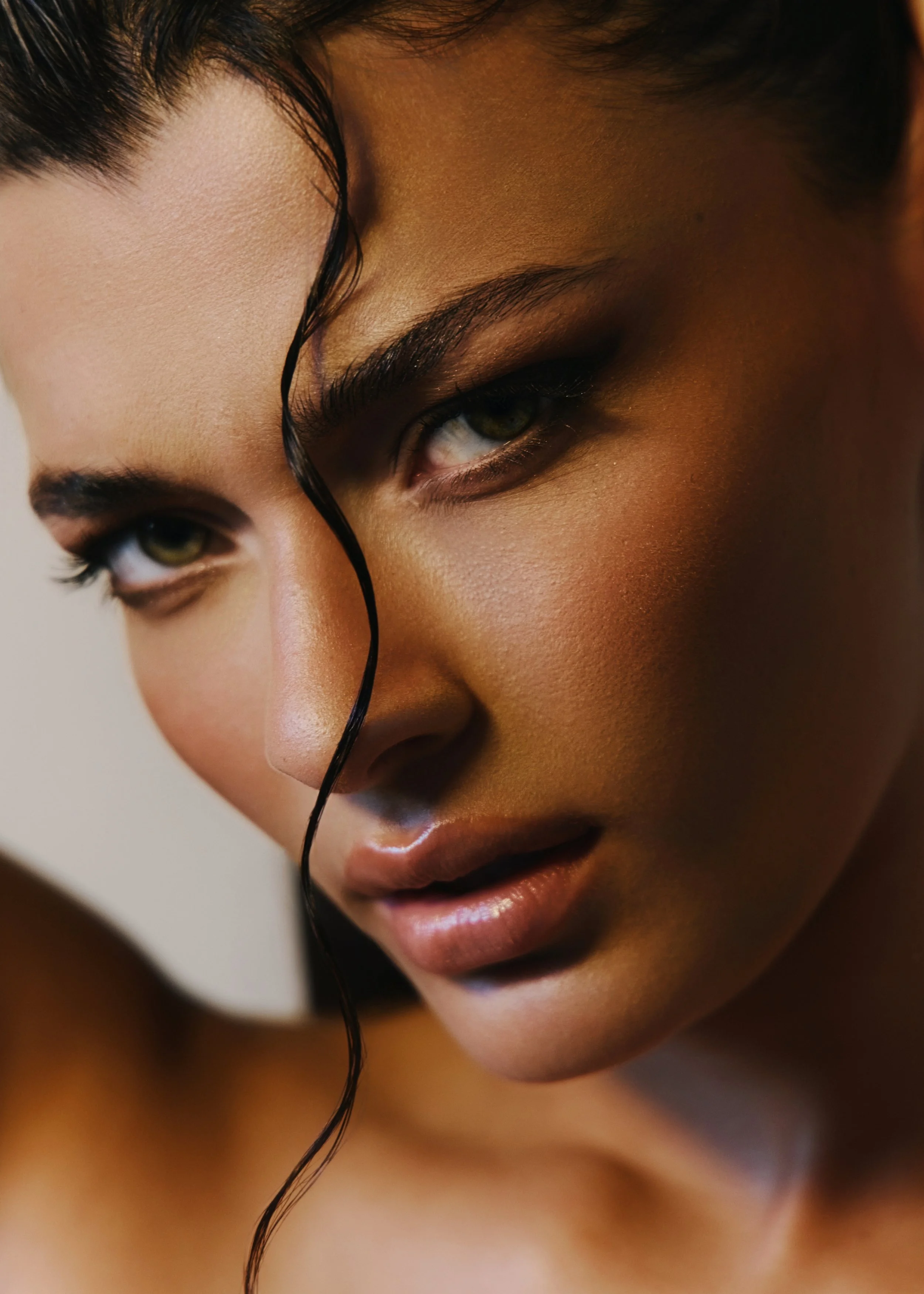 Close-up of a woman's face focusing on her eye, with a single black curl falling over her eyebrow, and natural makeup highlighting her features.