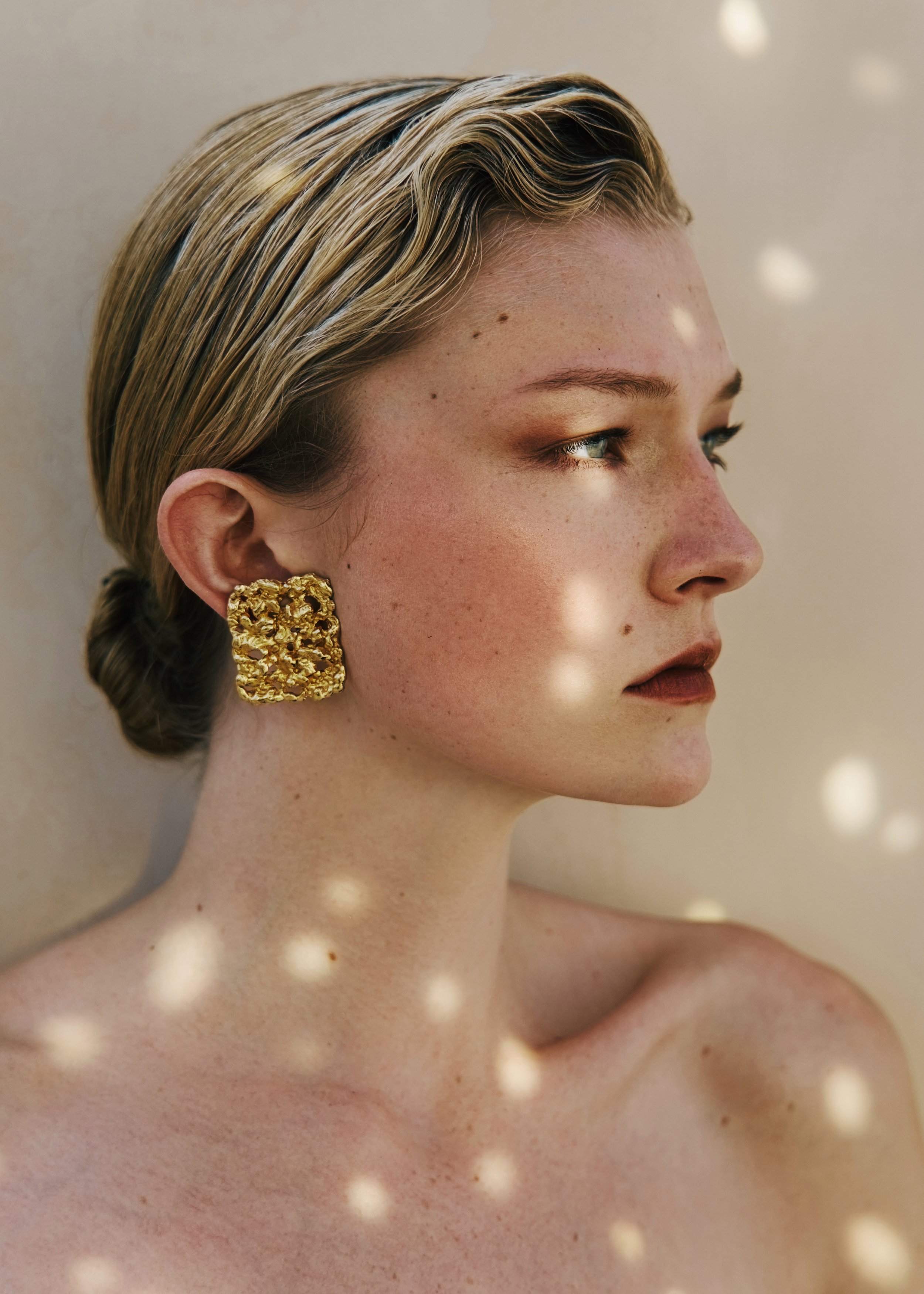 Close-up of a woman with blonde, wavy hair, wearing a large textured gold earring, and with freckles on her face and neck.