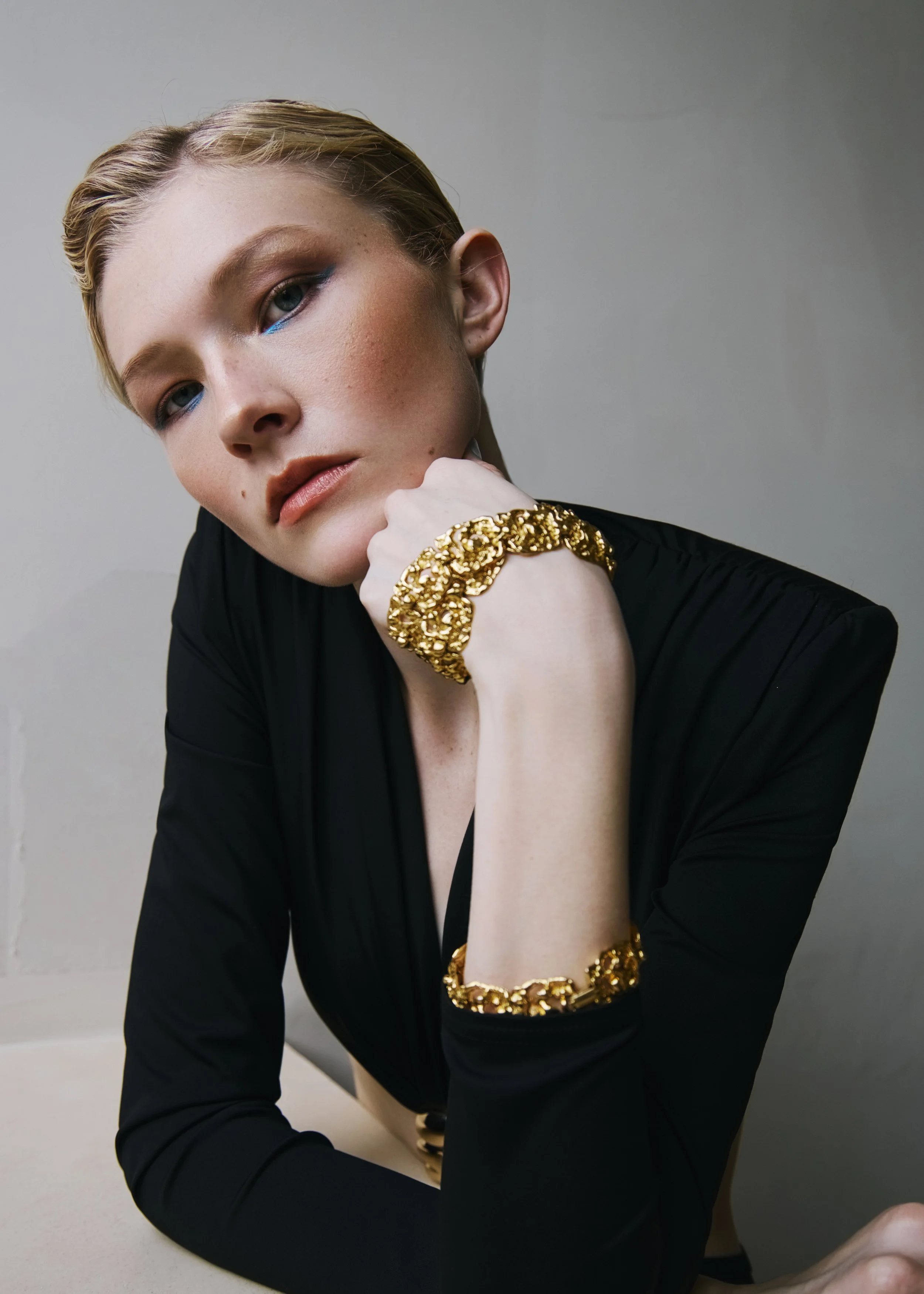 Close-up of a woman with short blonde hair, blue eyes, wearing blue eyeshadow, a black top, and gold jewelry, posing with her hand resting on her chin against a plain background.