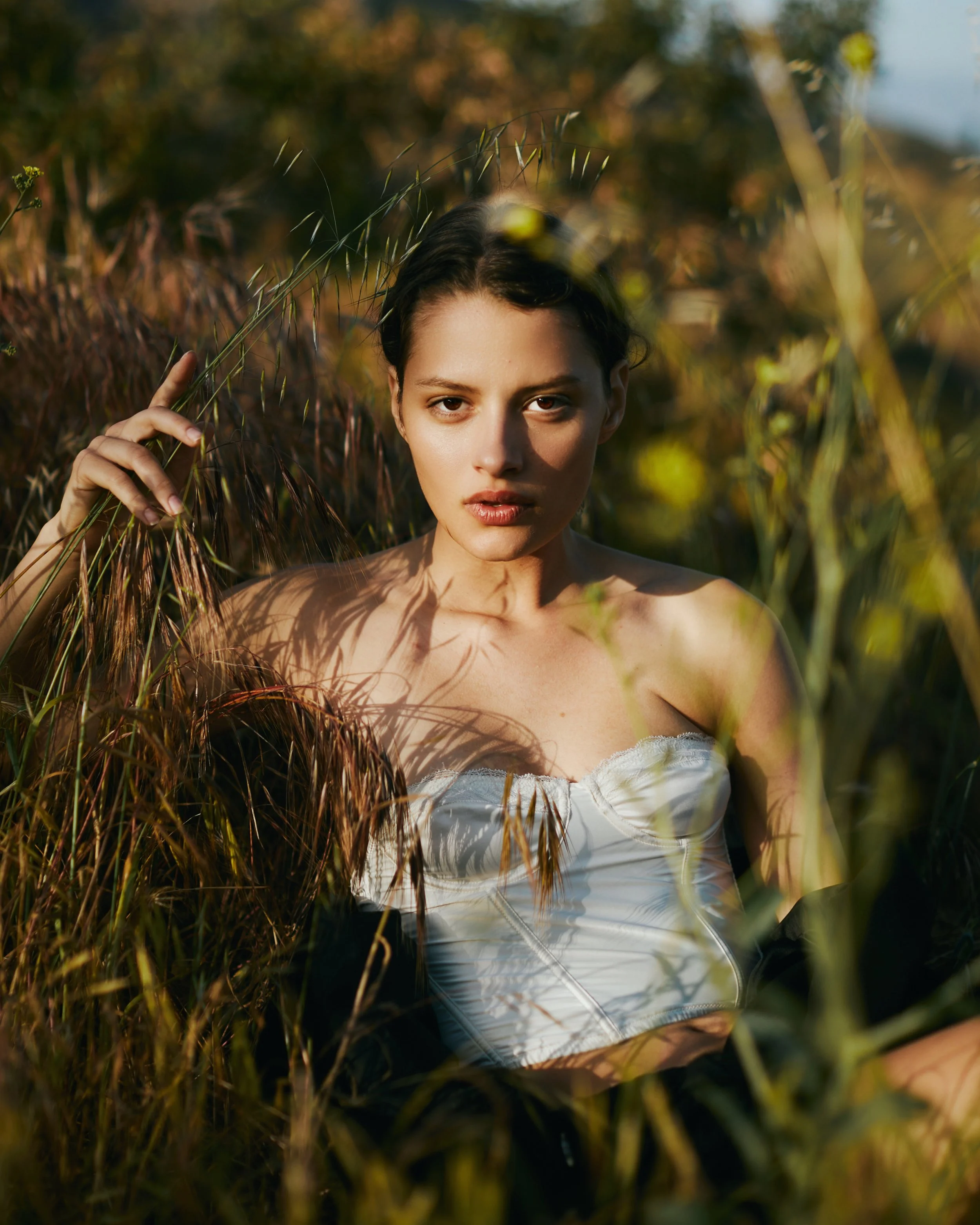 A woman with dark hair and a white blouse standing among tall grasses and plants outdoors during sunset.