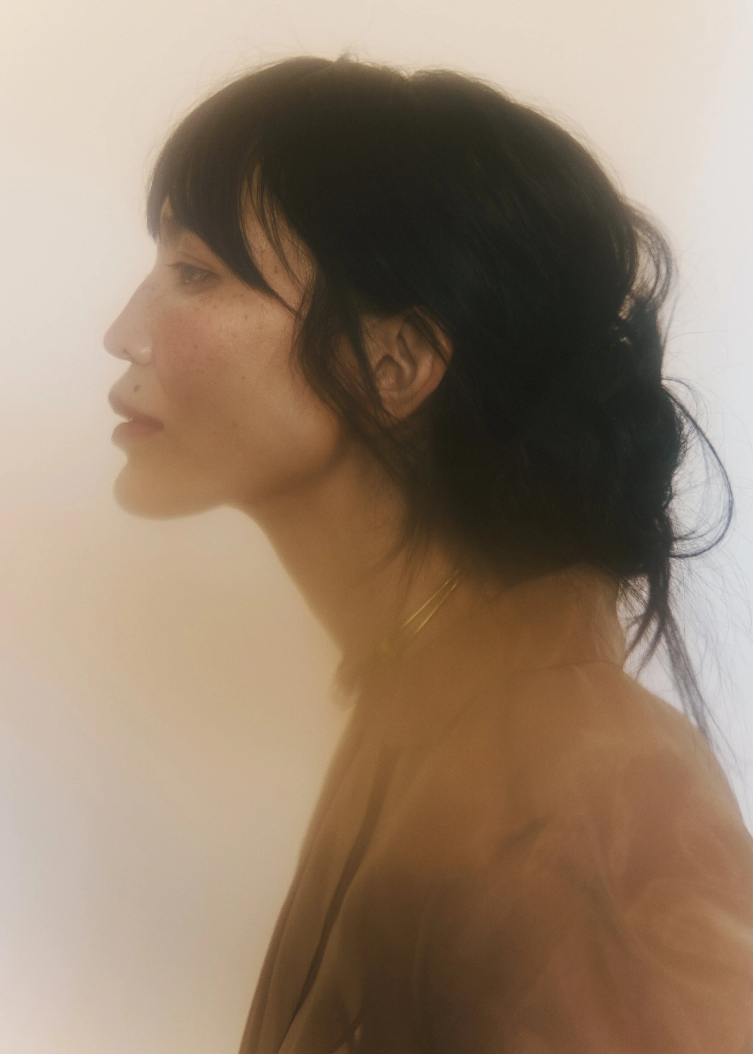 Side profile of a woman with dark hair, freckles, and a gold necklace, standing against a plain light-colored background.