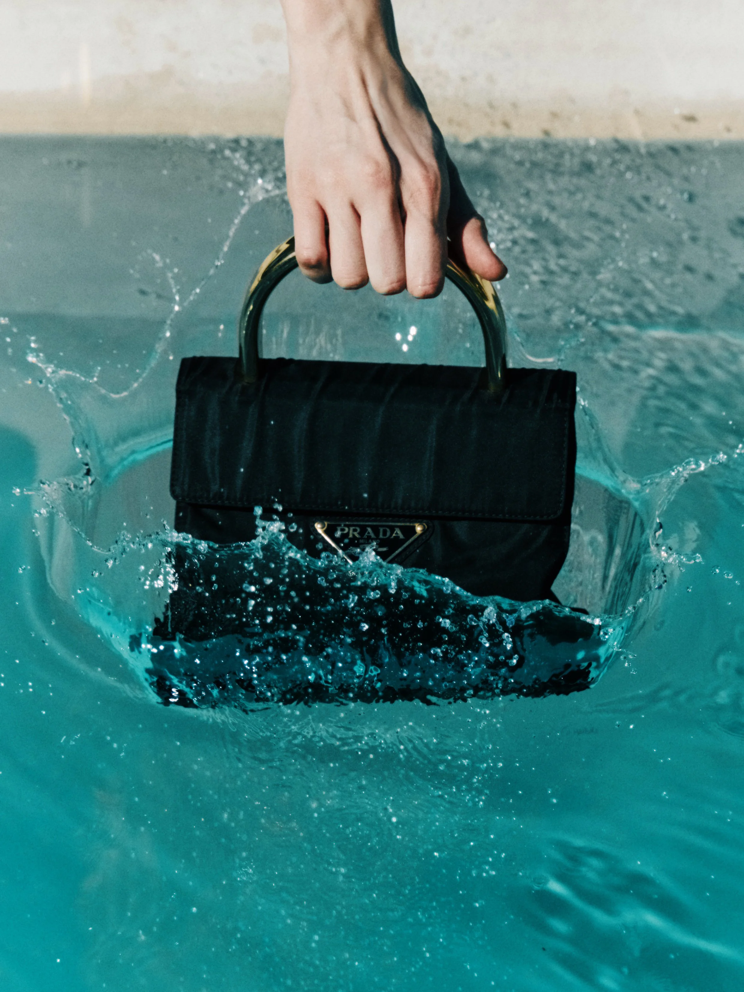Close-up of a person's hand gripping a Prada handbag handle as it emerges from the water, creating splash and ripples.